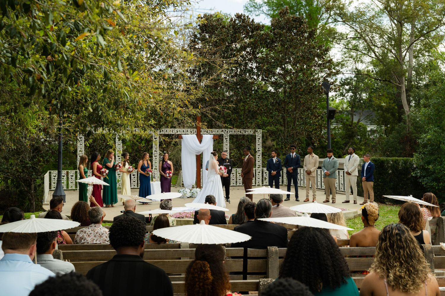 Outdoor wedding ceremony with guests seated on white chairs, bridal party standing at altar surrounded by lush greenery.