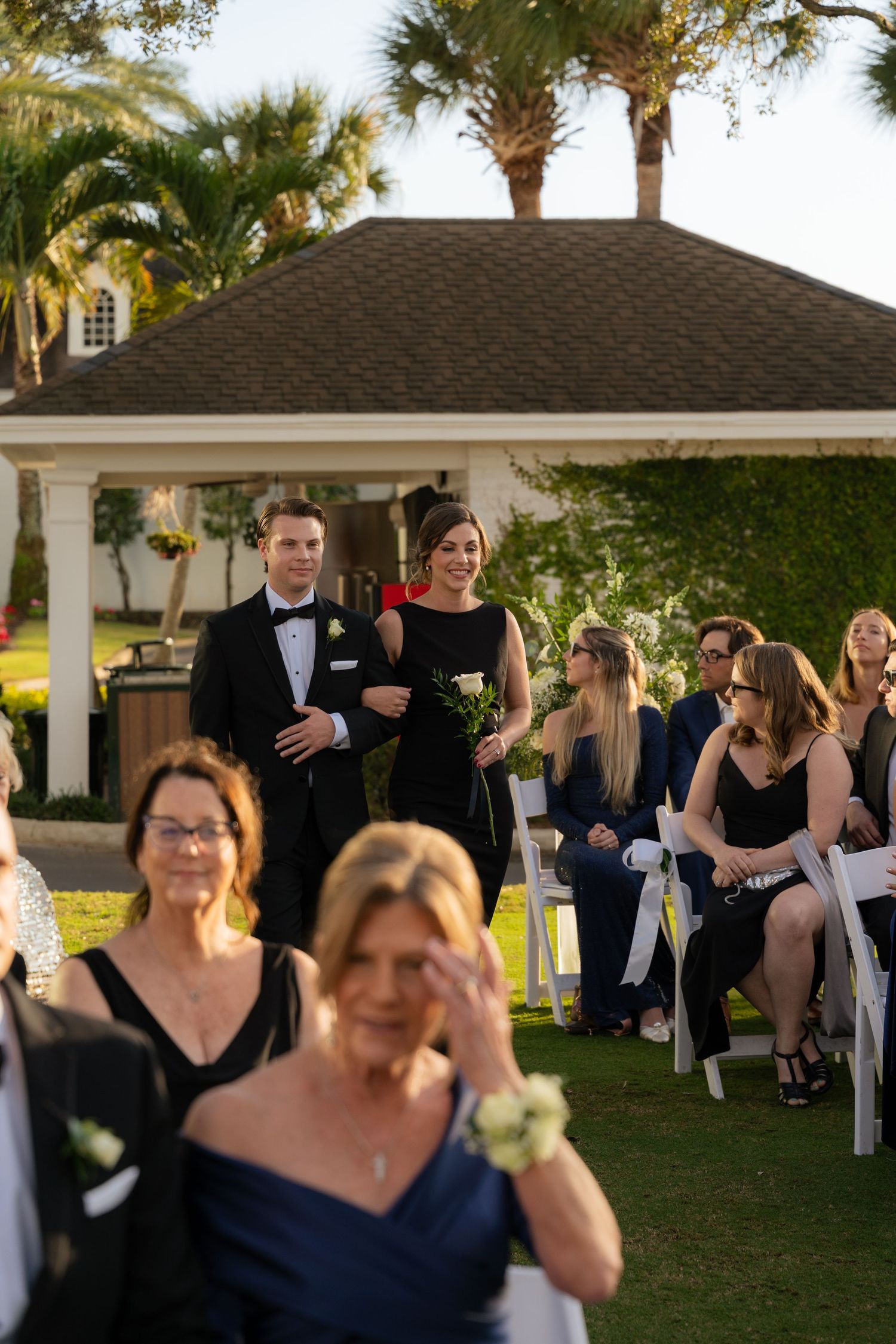 A groomsman escorts a bridesmaid down the aisle at an elegant outdoor wedding ceremony with palm trees.