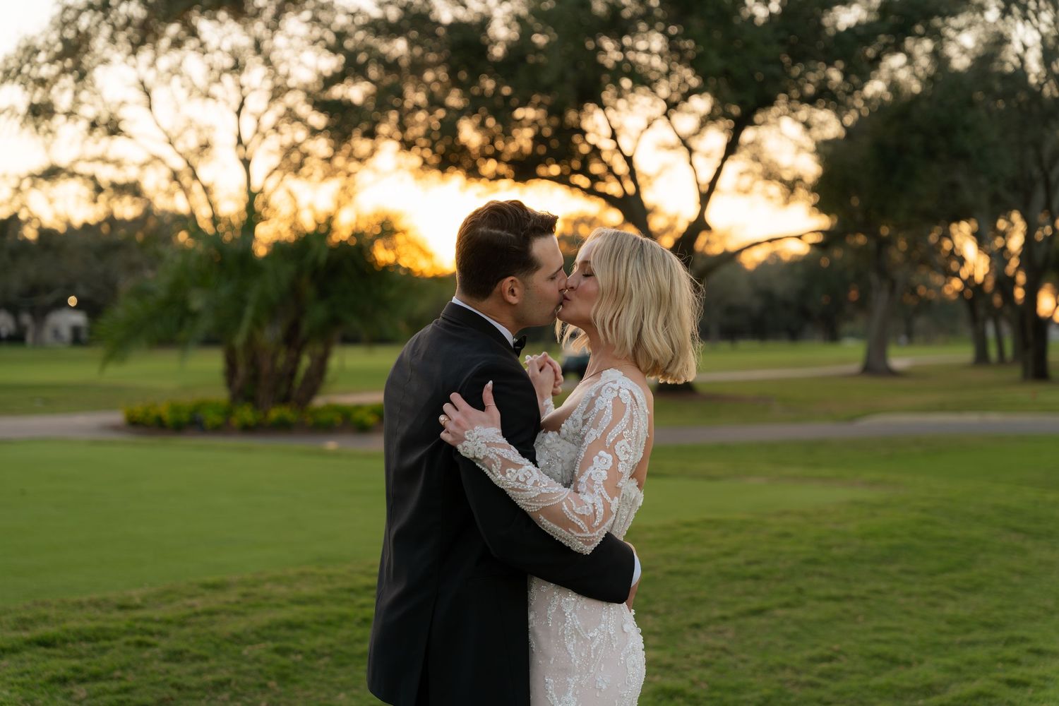 Bride and groom share romantic sunset kiss outdoors, bride in long-sleeve lace wedding gown on golf course.