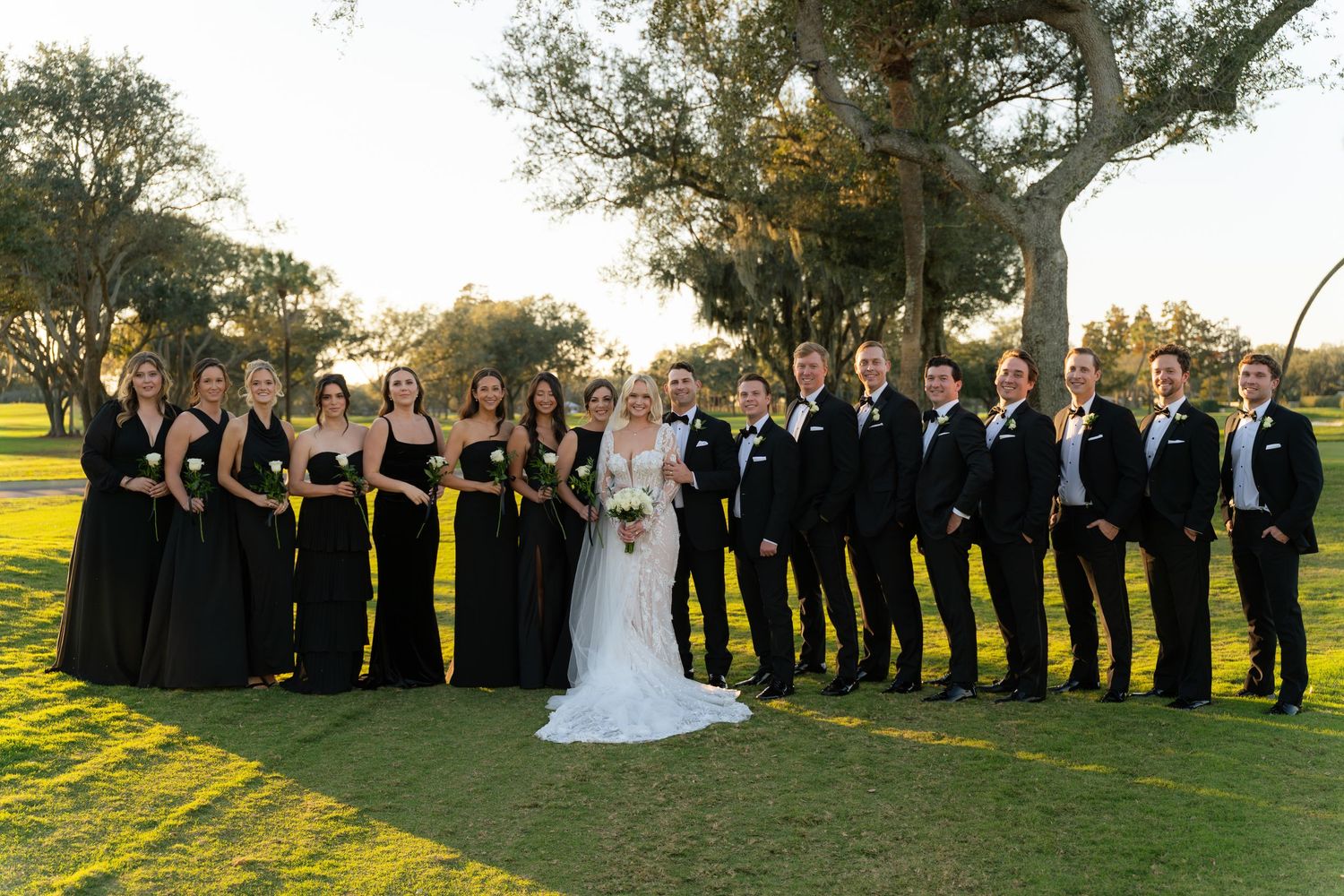 Wedding party in black attire poses outdoors on green lawn with trees, bride in white gown centered among bridesmaids and groomsmen.