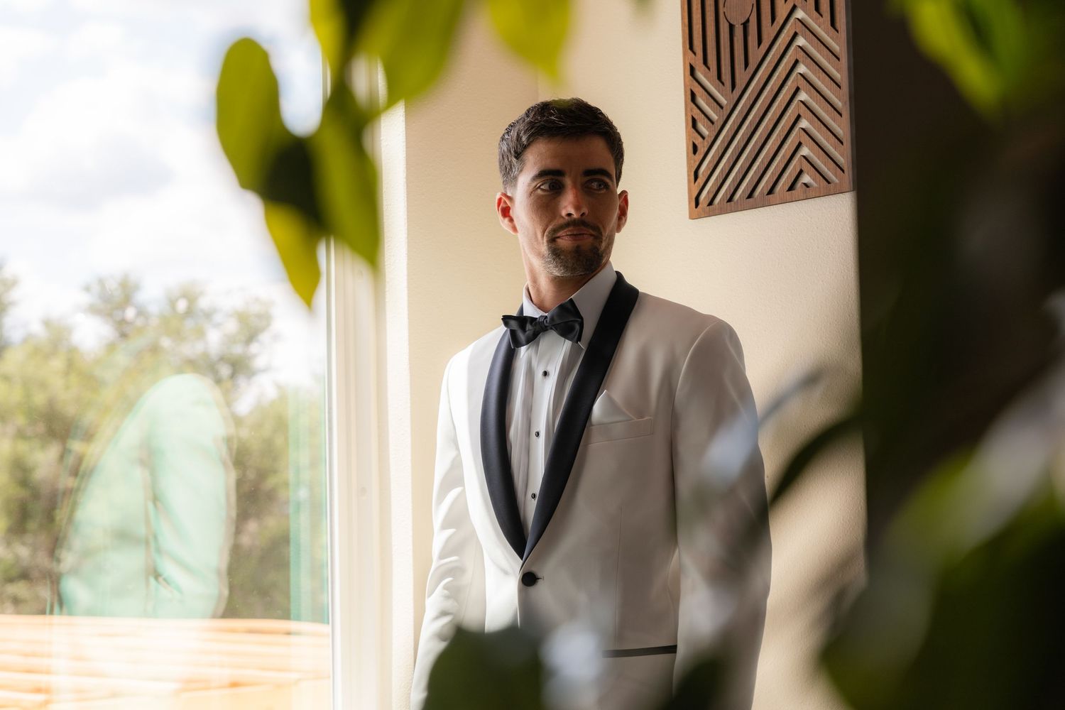 A young man in a white tuxedo with black bow tie stands elegantly by a window surrounded by lush green plants.