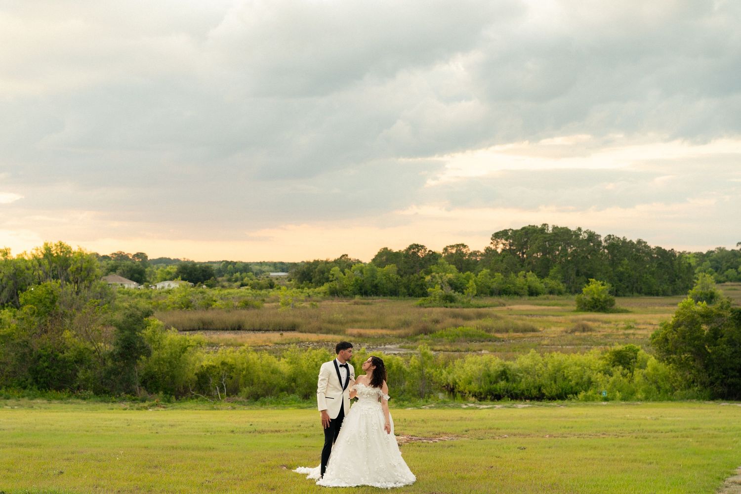 Bride and groom stand together in a vast green meadow under a dramatic cloudy sky at sunset.