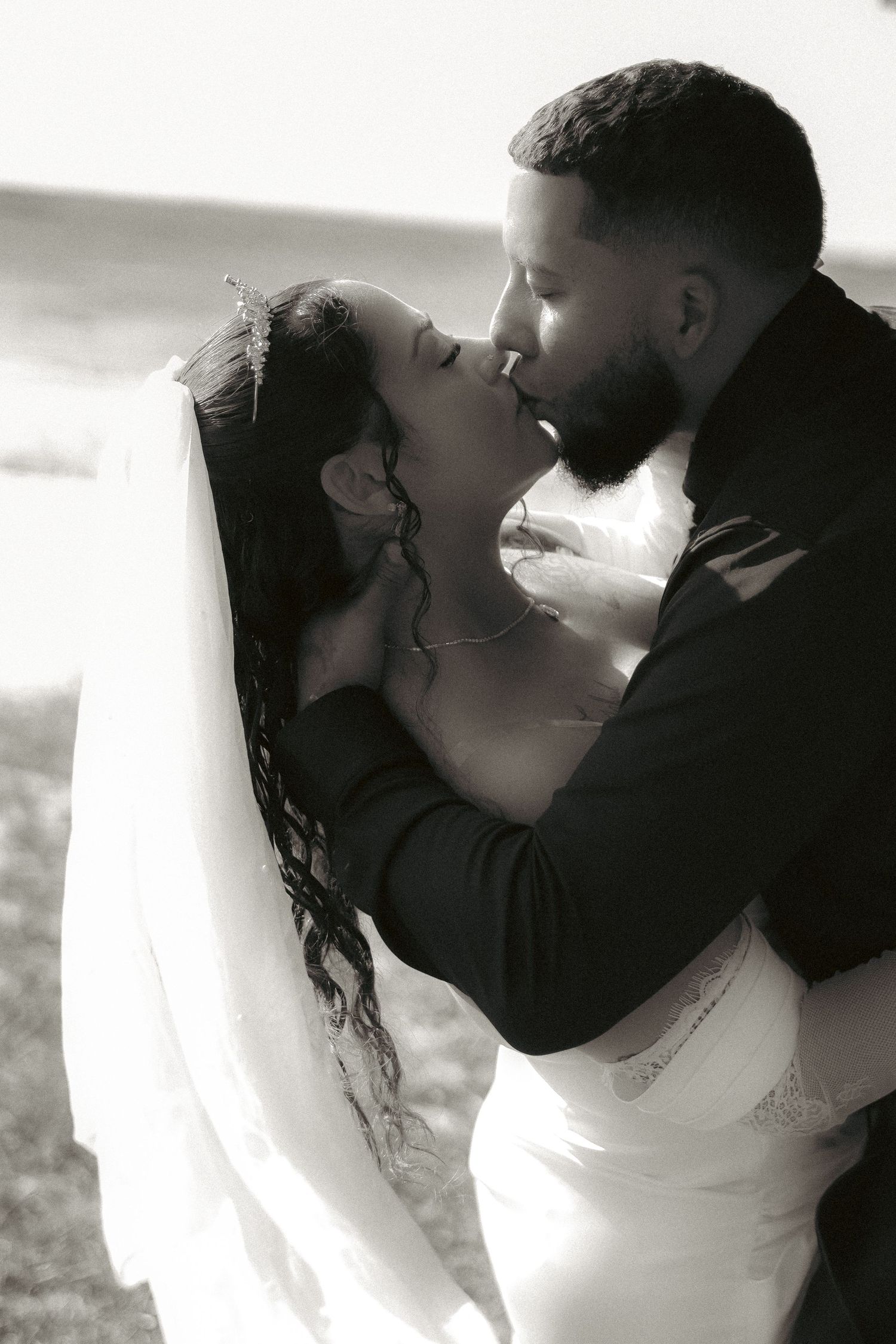 Black and white photo of a bride and groom sharing a romantic kiss on their wedding day outdoors.