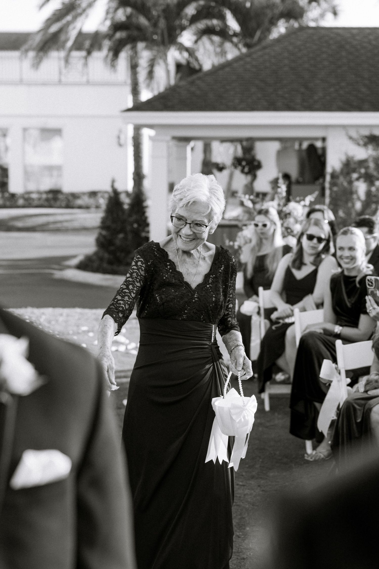 Elderly woman in black lace dress walks down outdoor wedding aisle carrying white flower basket.