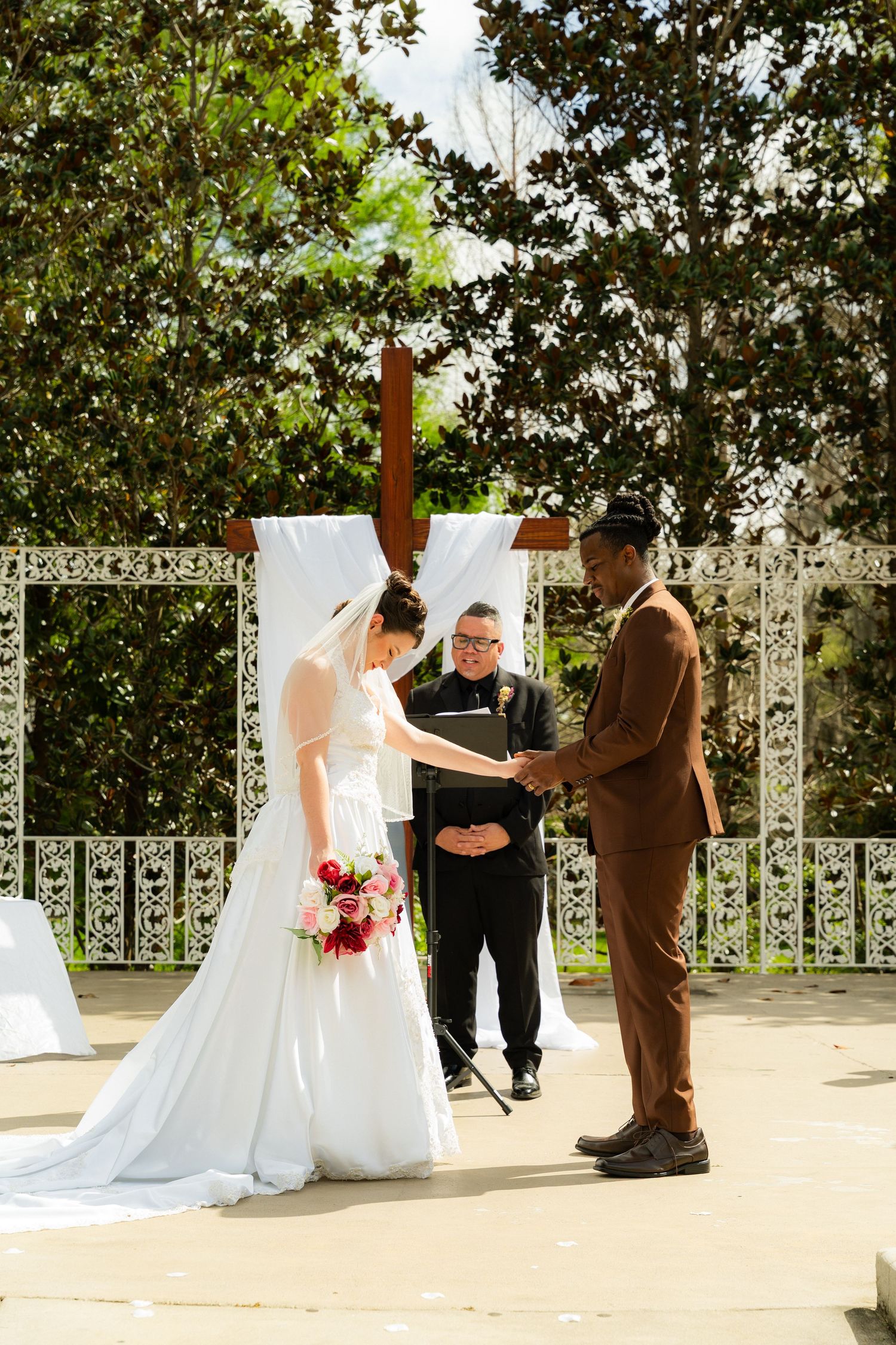 Bride and groom hold hands at outdoor wedding altar with cross backdrop as officiant presides over the ceremony.
