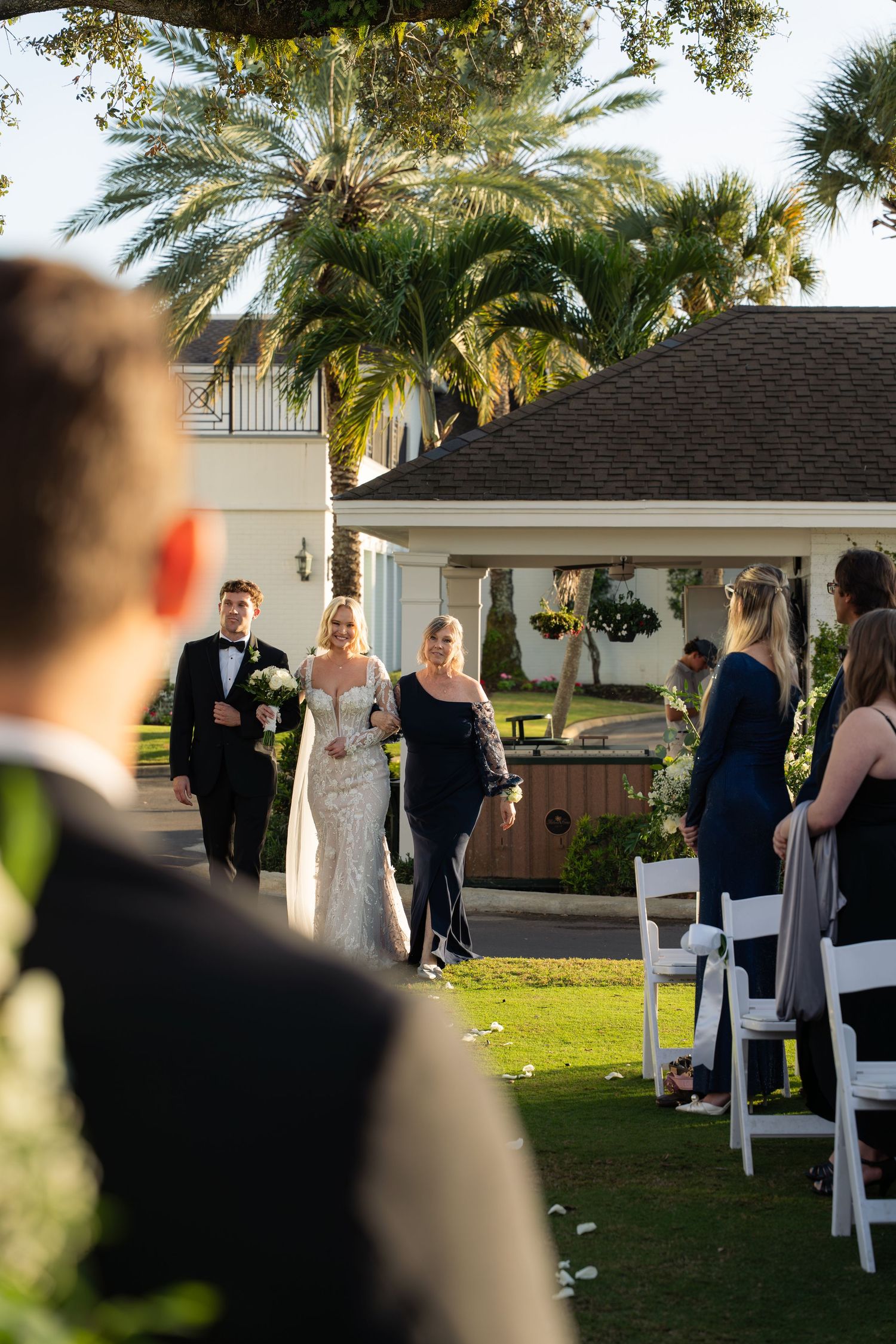 Bride in white gown walks down outdoor aisle with escorts at sunny Florida wedding ceremony.