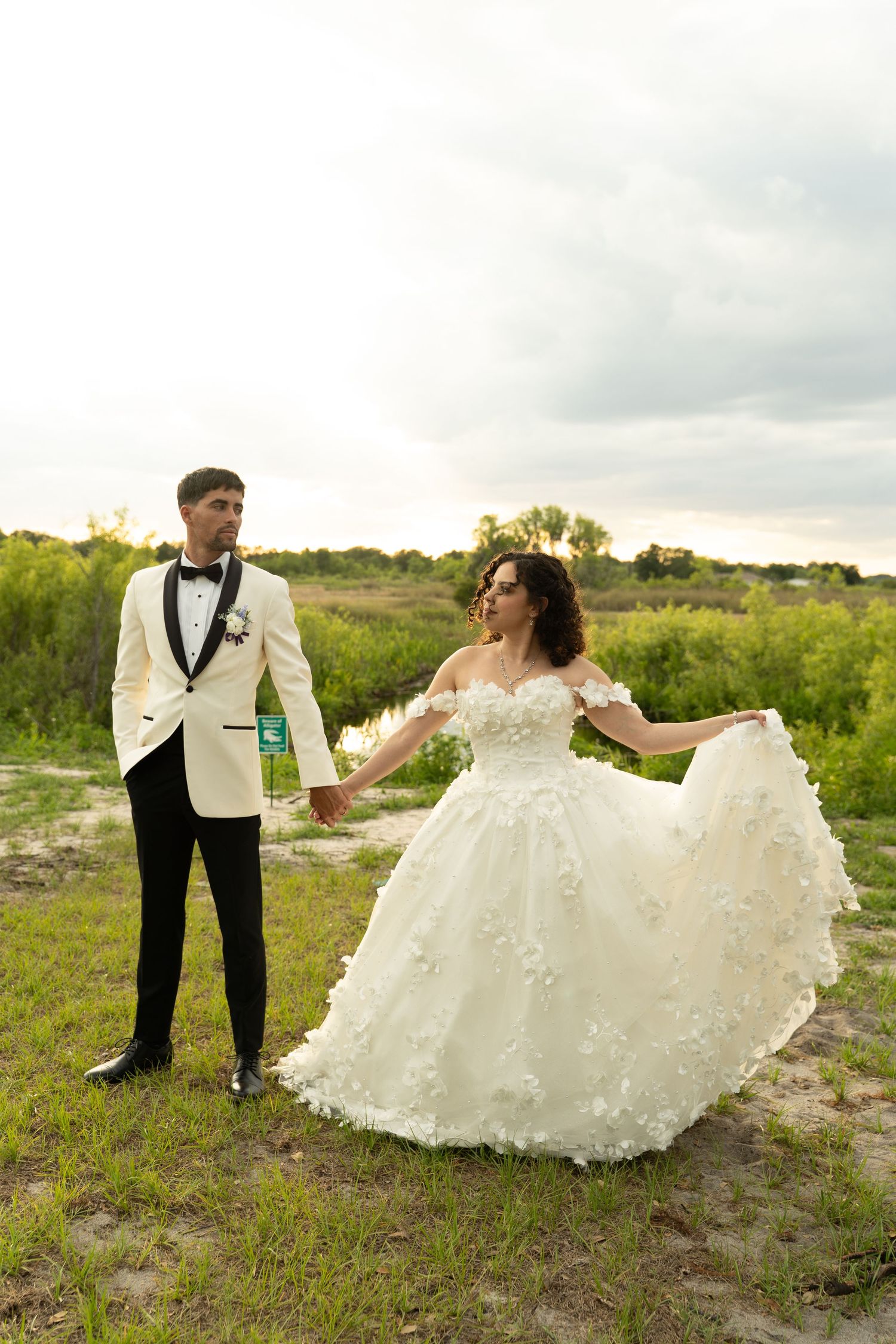Bride in off-shoulder floral wedding gown holding hands with groom in white tuxedo outdoors.