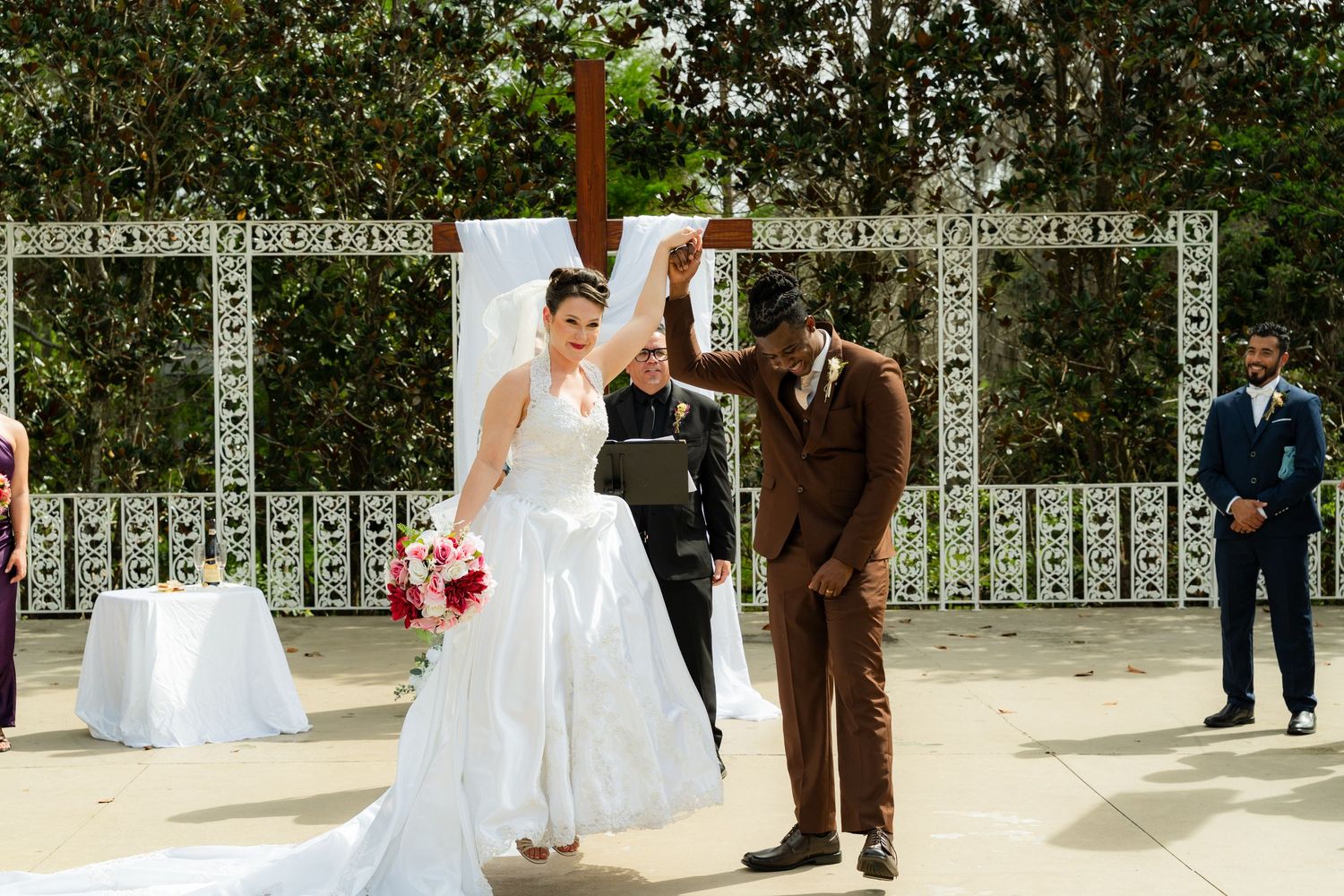Bride and groom celebrate at outdoor wedding ceremony with white lattice backdrop and wooden cross.