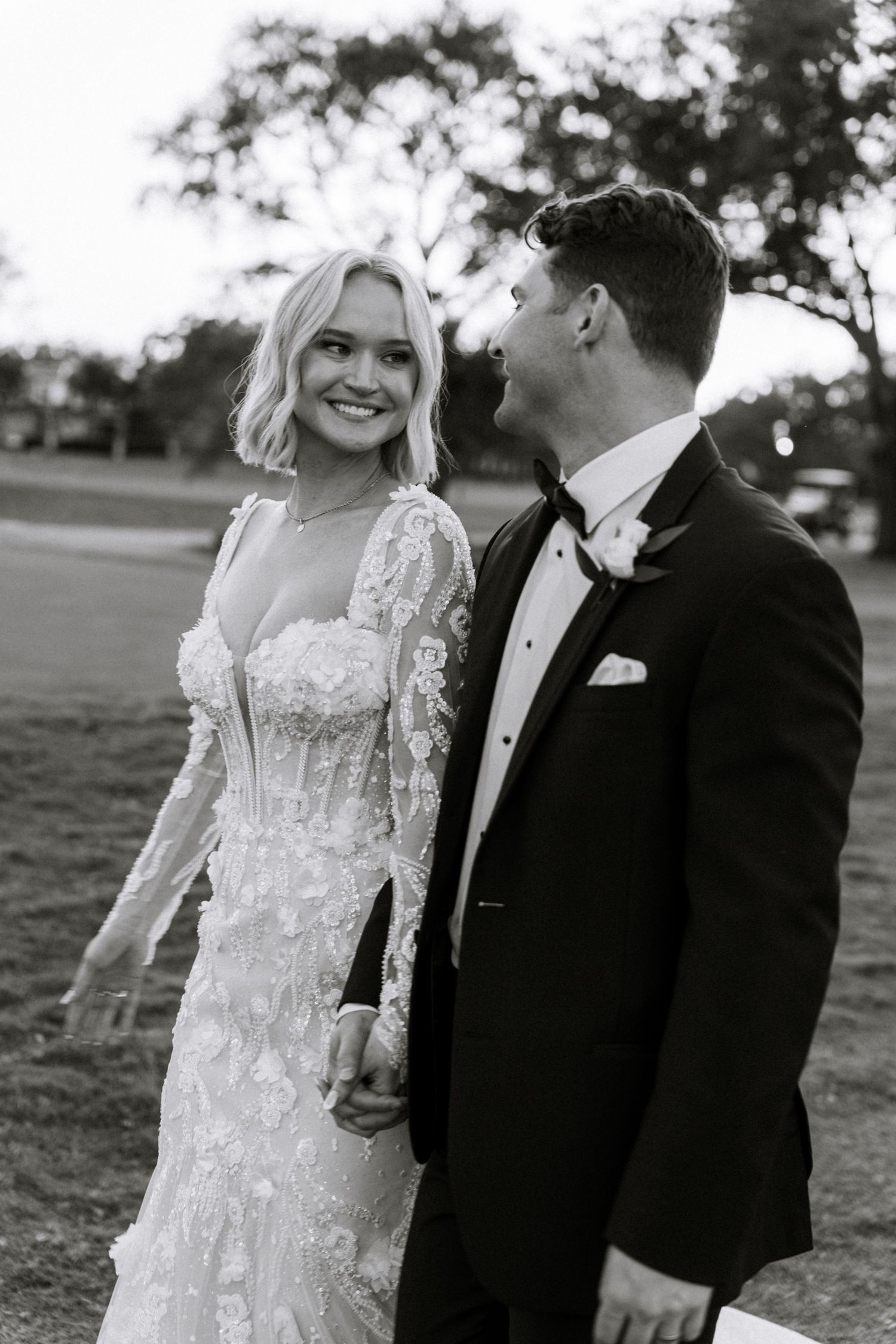 Black and white photo of a smiling bride in lace gown holding hands with groom in tuxedo outdoors.