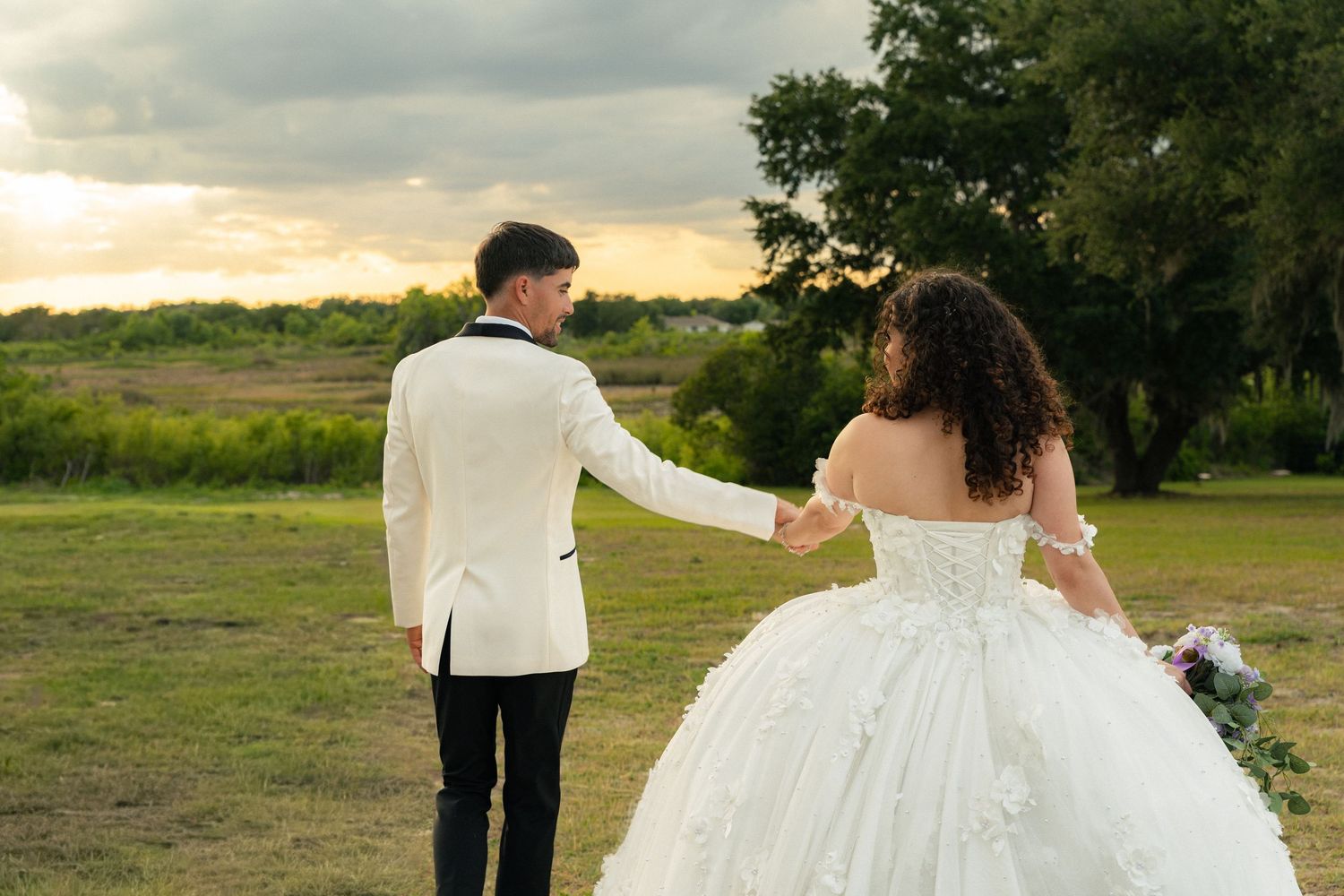Bride and groom walking hand in hand across green field at sunset, bride in ball gown wedding dress.