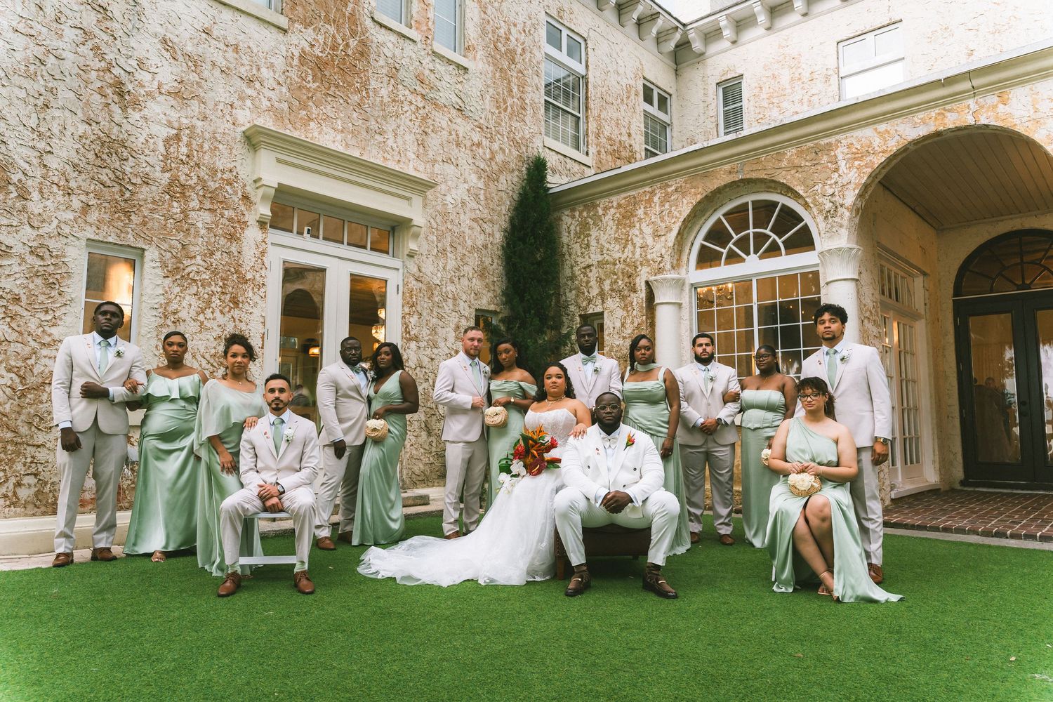 Wedding party poses outside elegant stone mansion, bridesmaids in mint green gowns and groomsmen in gray suits flanking bride and groom.