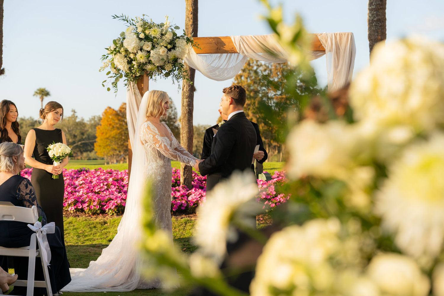 Outdoor wedding ceremony with bride and groom at floral arch, white blooms in foreground, pink flowers and palm trees behind.