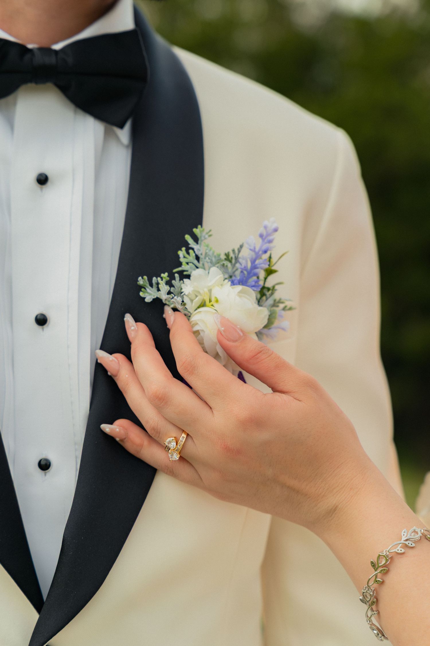 Bride adjusting white and blue floral boutonniere on grooms ivory tuxedo lapel, showcasing wedding ring and bracelet.