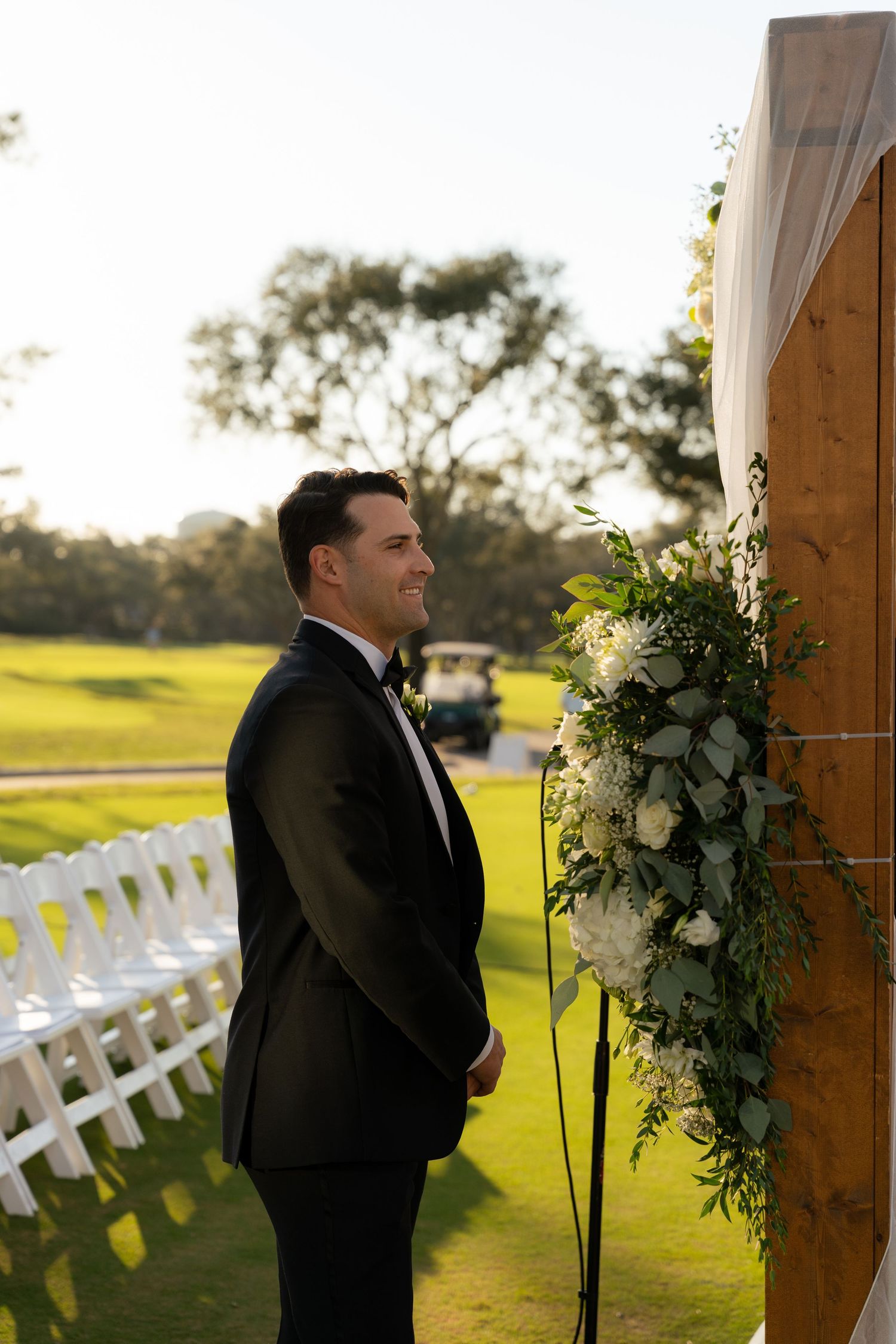 Groom in black tuxedo smiling at outdoor wedding ceremony beside floral arch on lush green golf course.