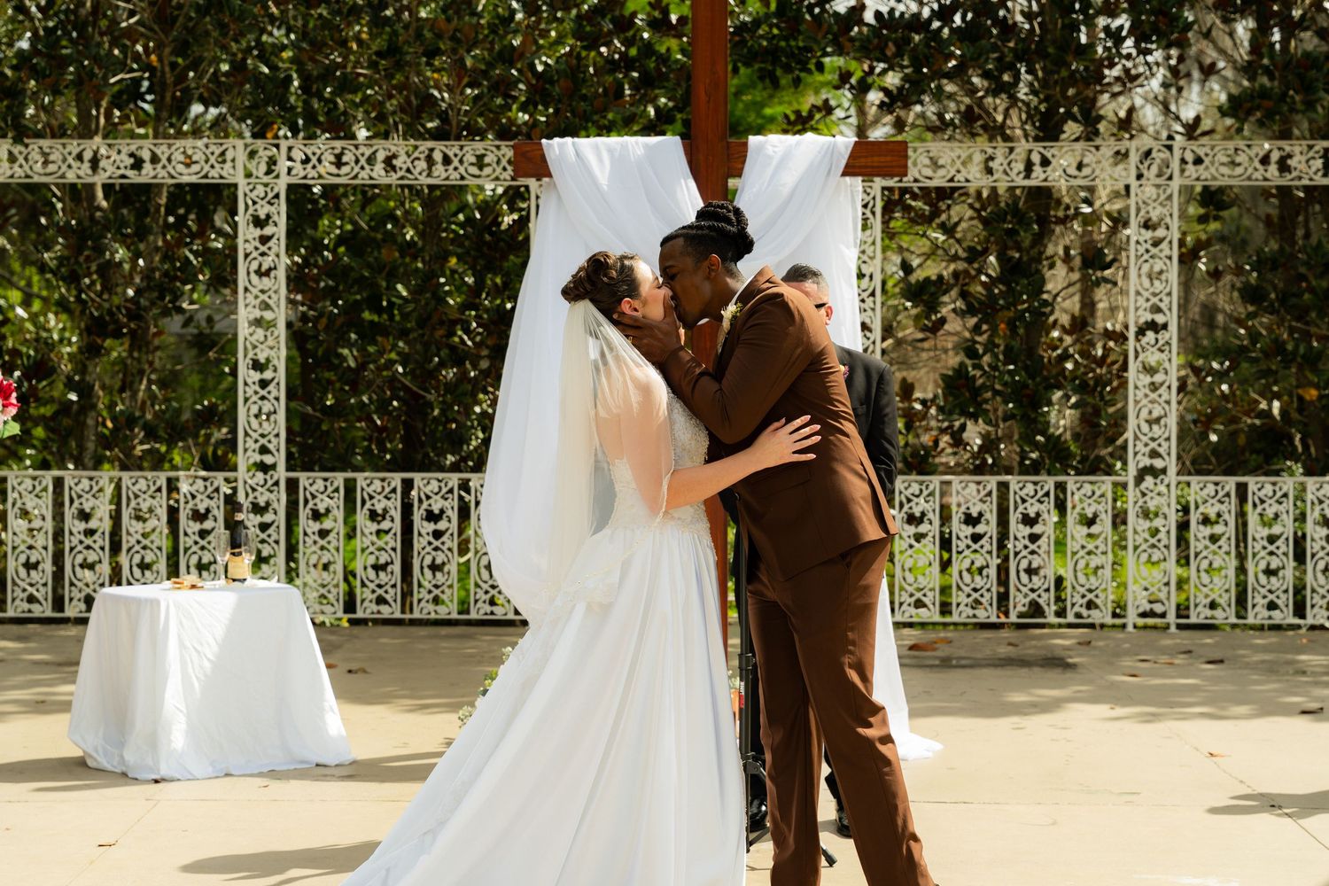 Bride and groom share first kiss at outdoor wedding ceremony beneath wooden cross with white draping.