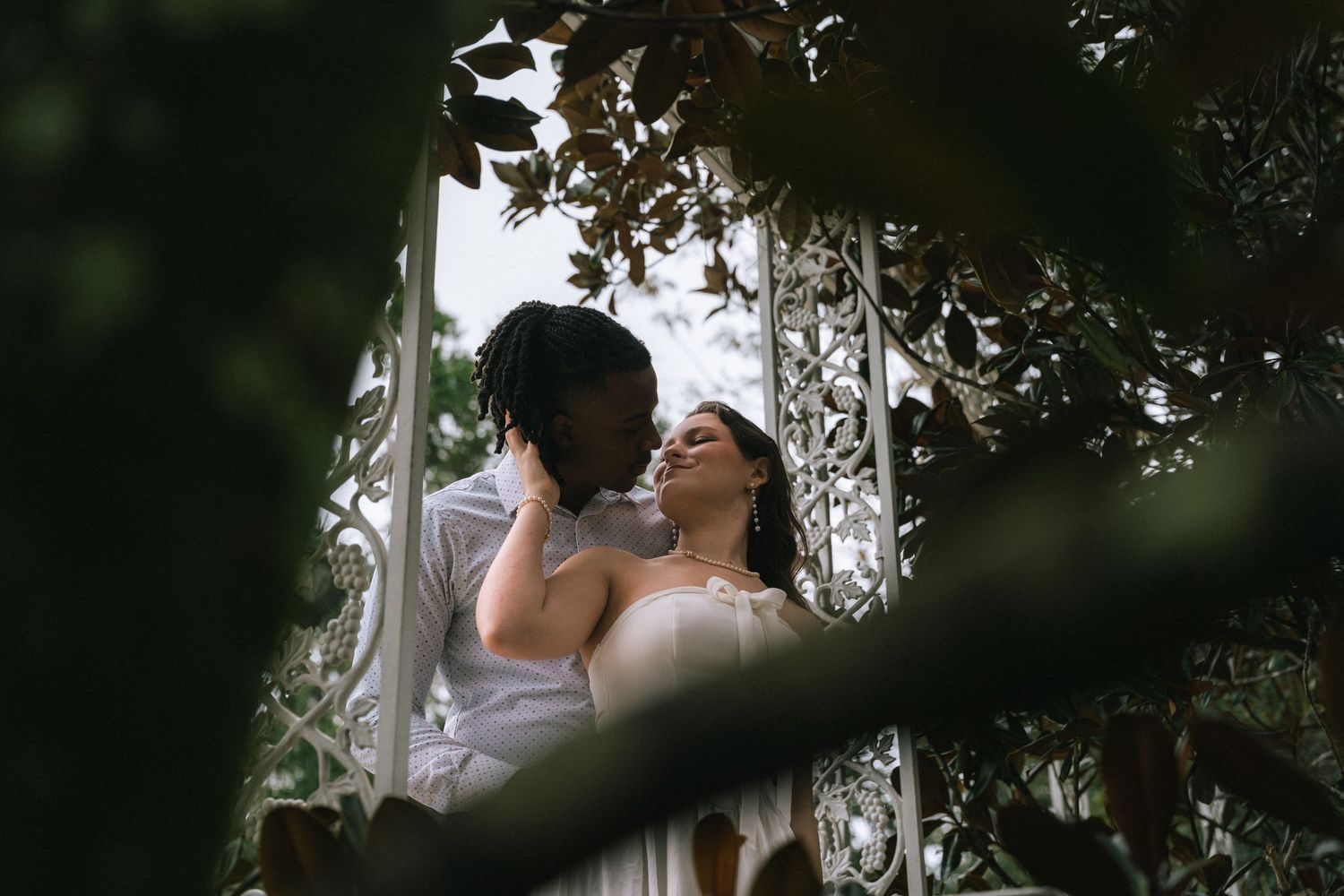 Couple sharing an intimate kiss under a canopy of trees and natural light during an outdoor wedding ceremony.