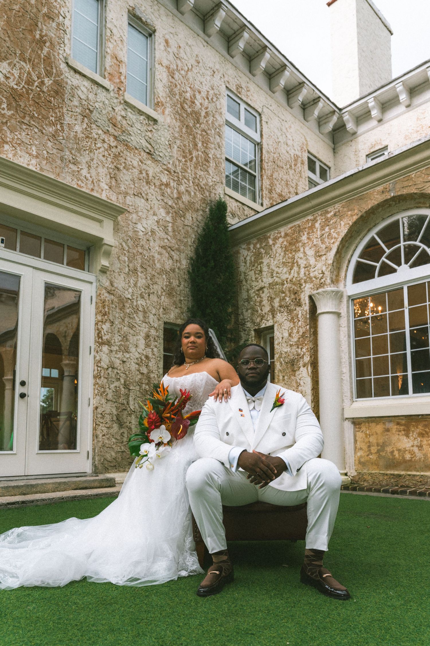 A bride and groom pose elegantly outside a historic stone mansion, she in a white gown with bouquet, he in a white suit.