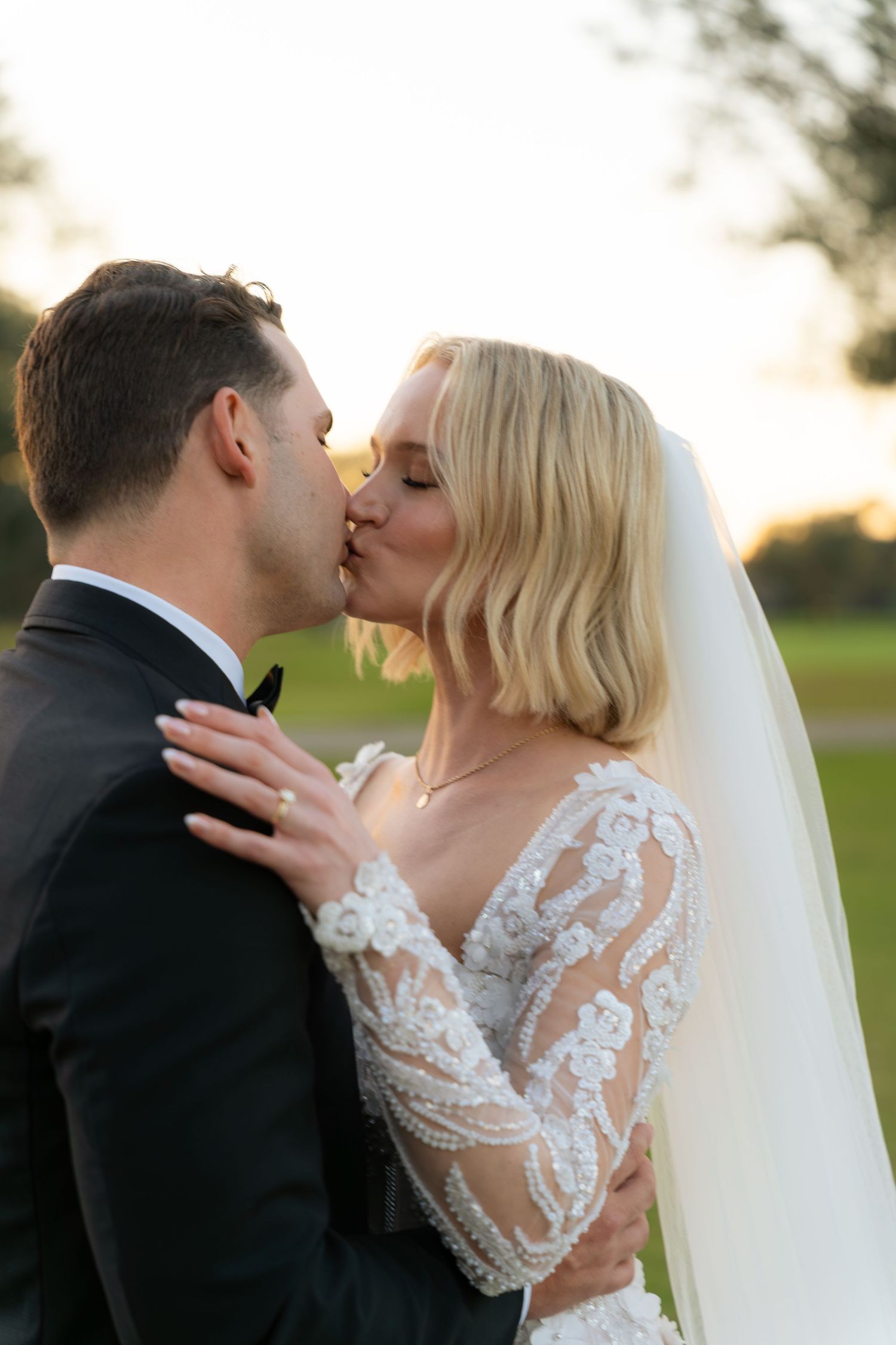 Bride and groom share a tender kiss at sunset, bride in long-sleeve lace gown with veil.