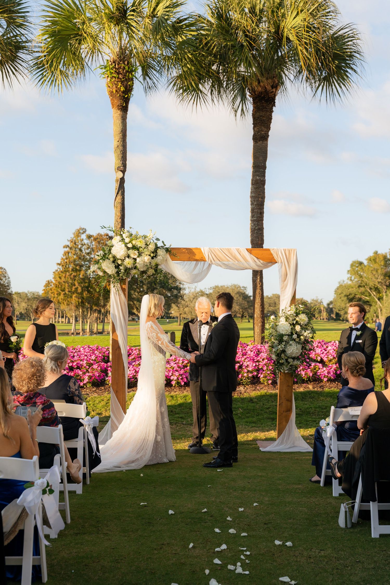 Bride and groom exchange vows under a floral arch flanked by palm trees at an outdoor golf course wedding ceremony.