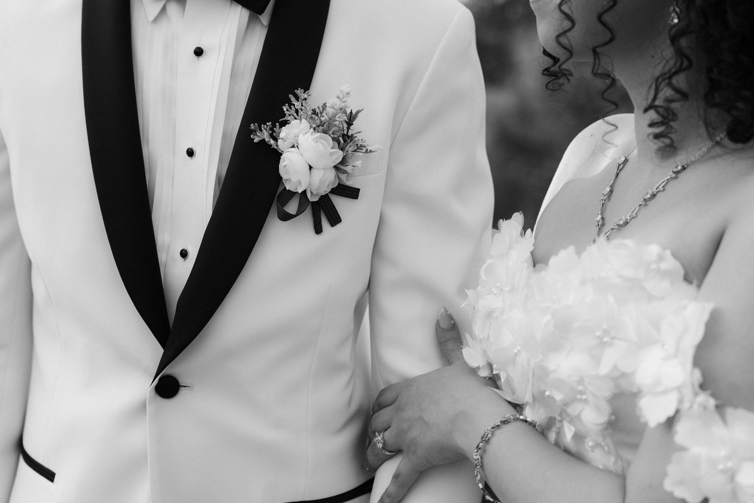 Black and white close-up of groom in white tuxedo with boutonniere standing beside bride in wedding dress.