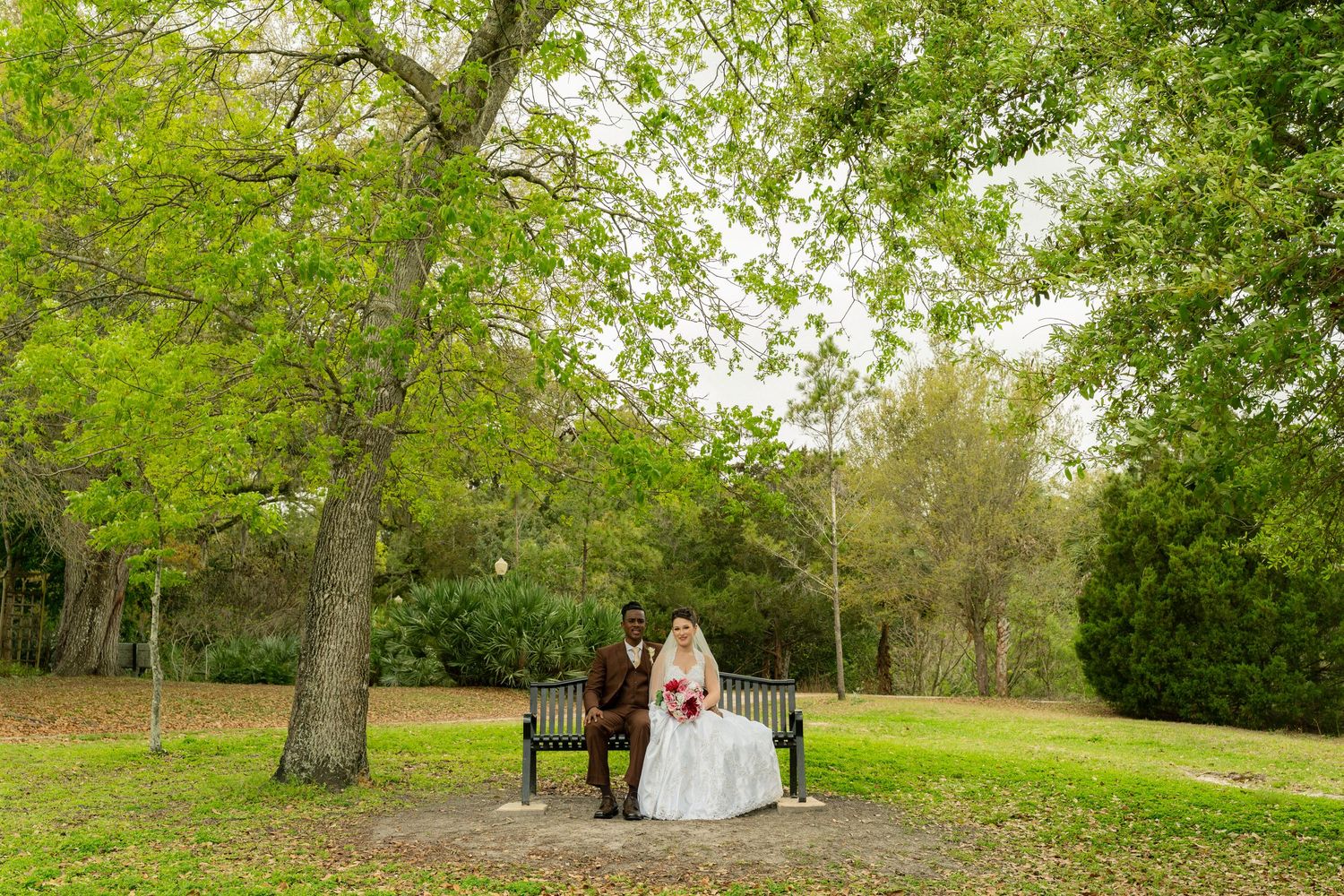 Bride and groom sitting on a park bench under a large green tree on their wedding day.