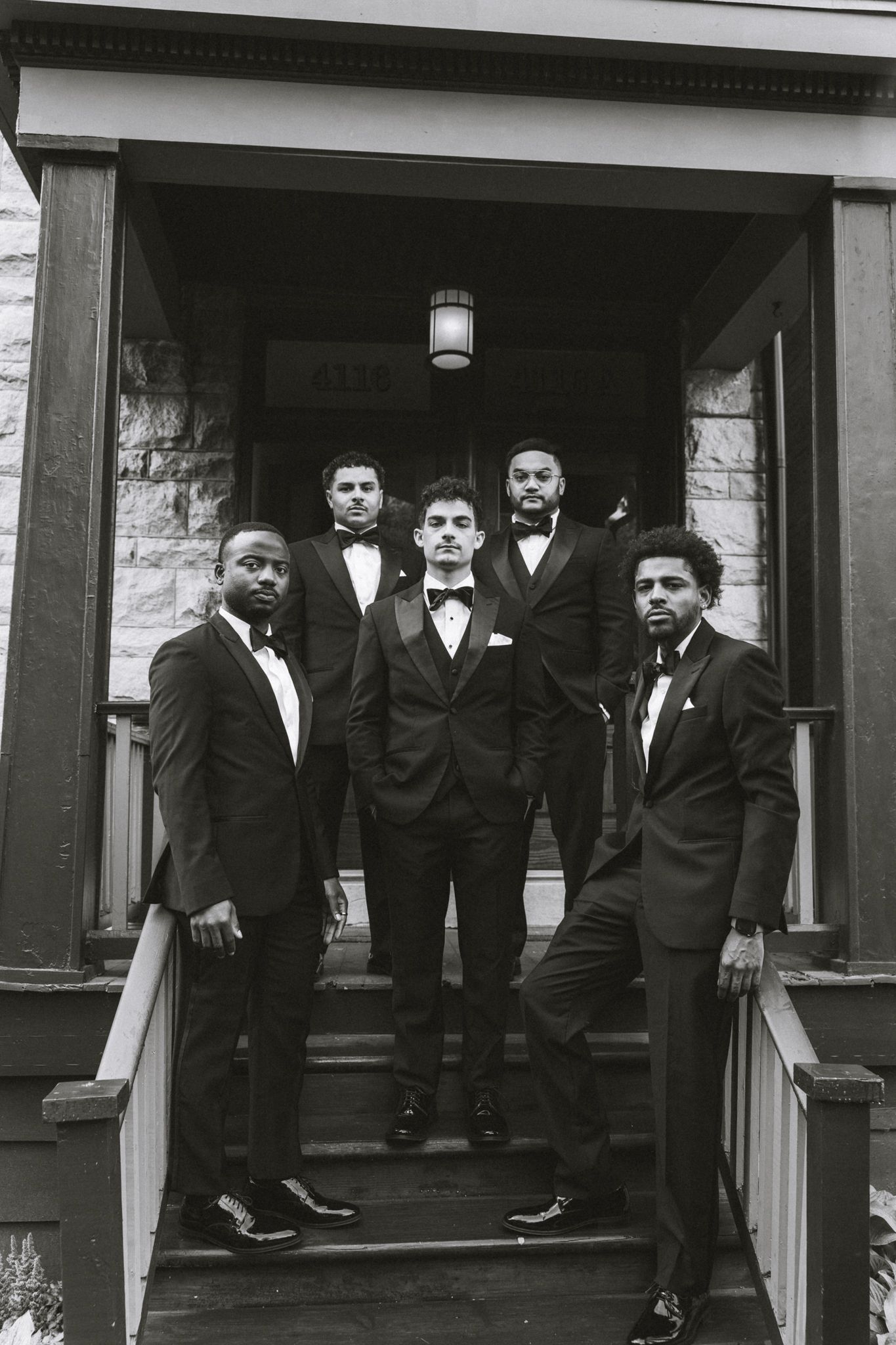 Five groomsmen in black tuxedos and bow ties pose on stone building steps in elegant black and white wedding photo.