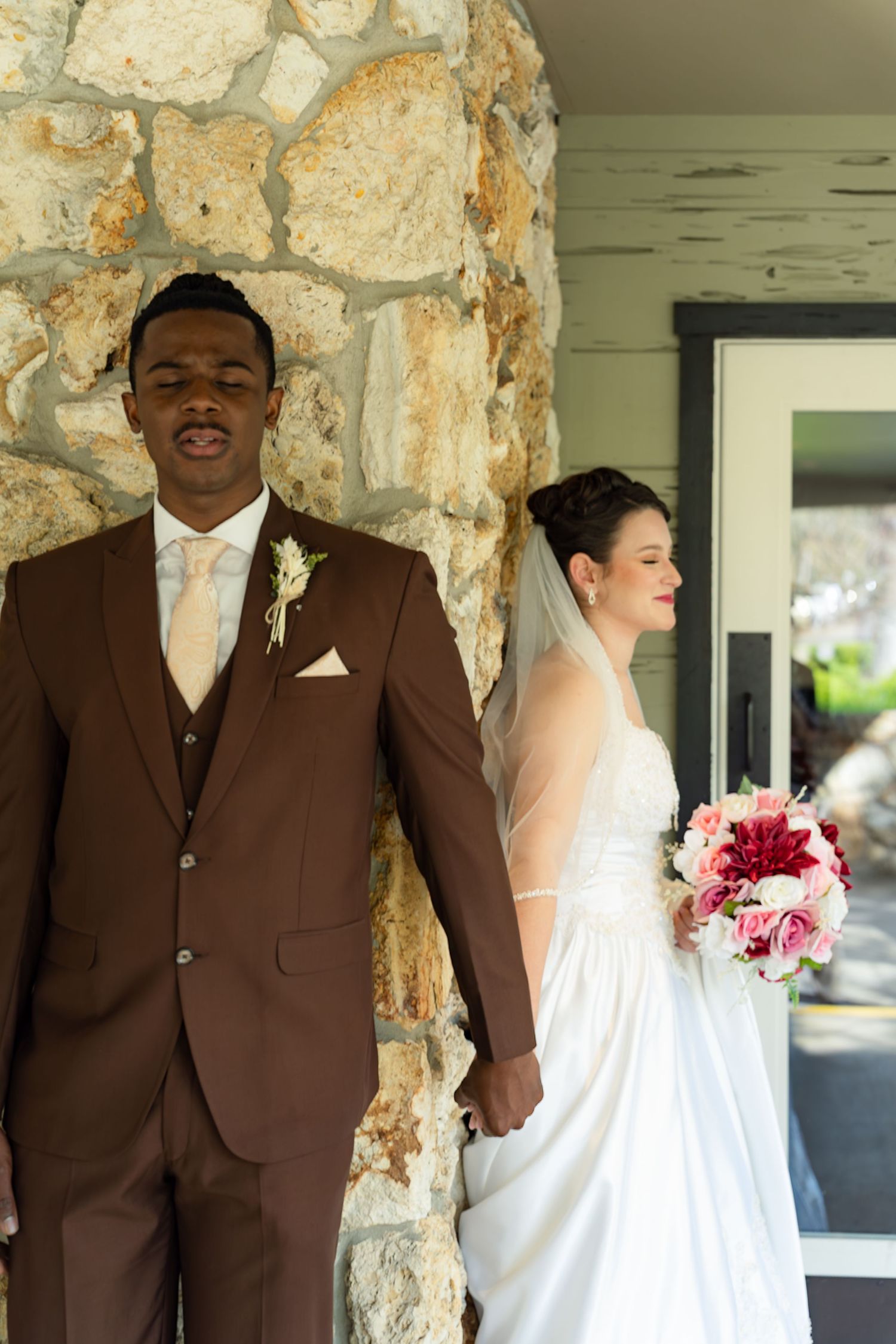 Interracial newlyweds hold hands by stone column, groom in brown suit, bride in white dress with veil and floral bouquet.