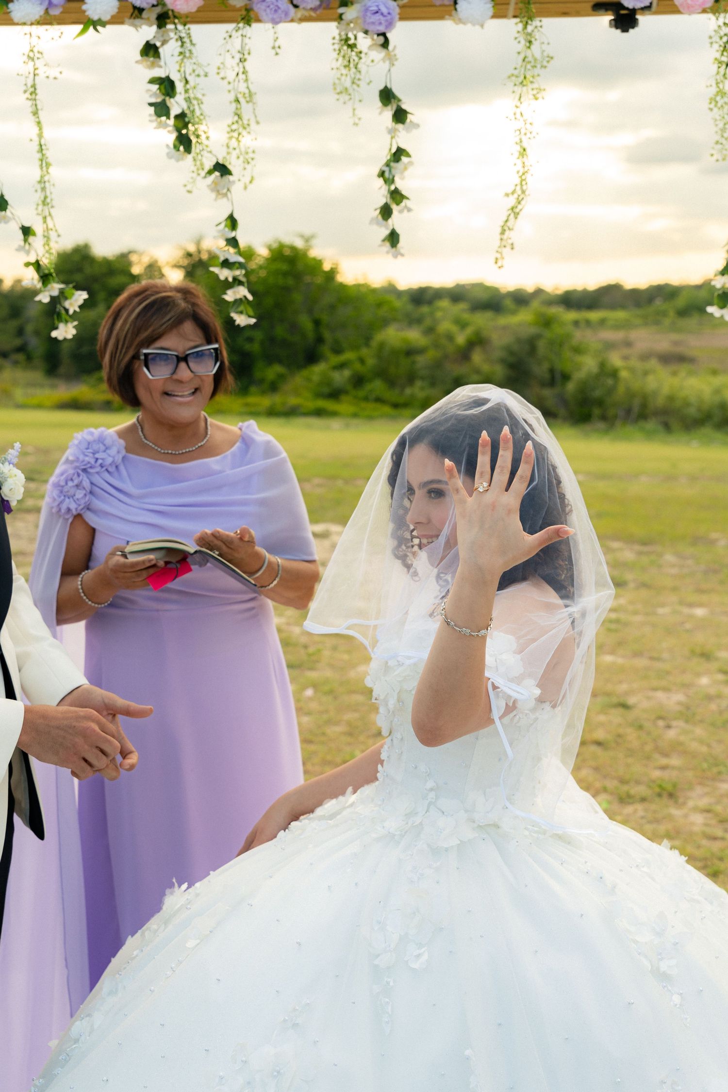 Bride in ball gown and veil shows her ring hand during outdoor wedding ceremony at sunset.