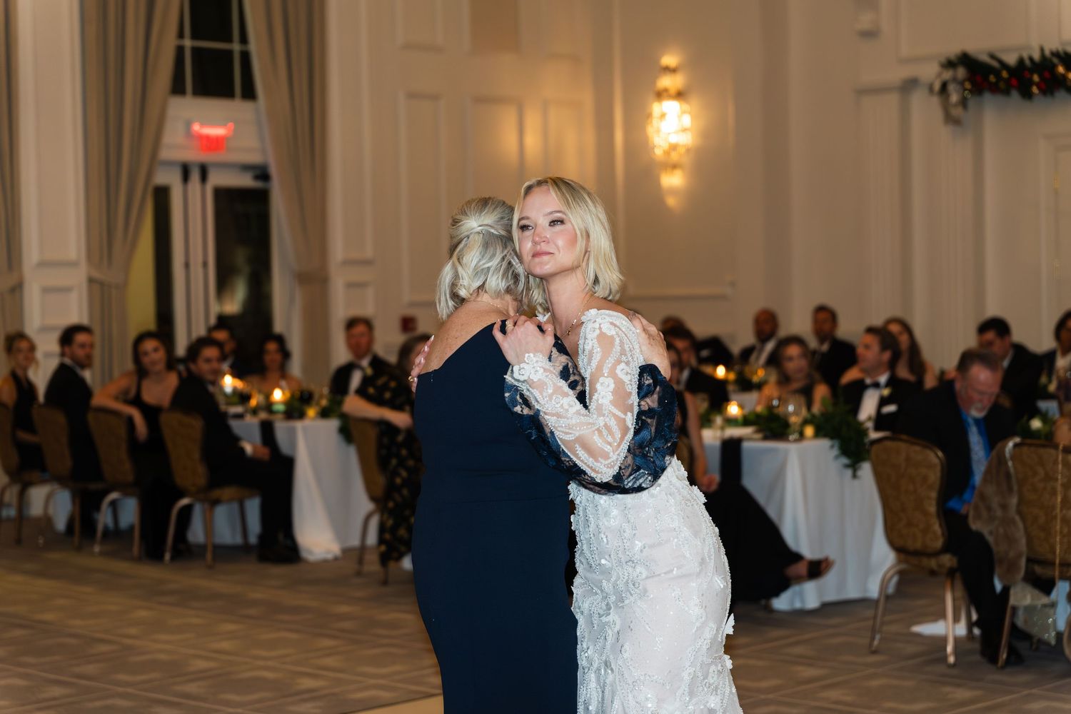 A bride in a lace gown shares an emotional dance with a guest in an elegant candlelit ballroom reception.