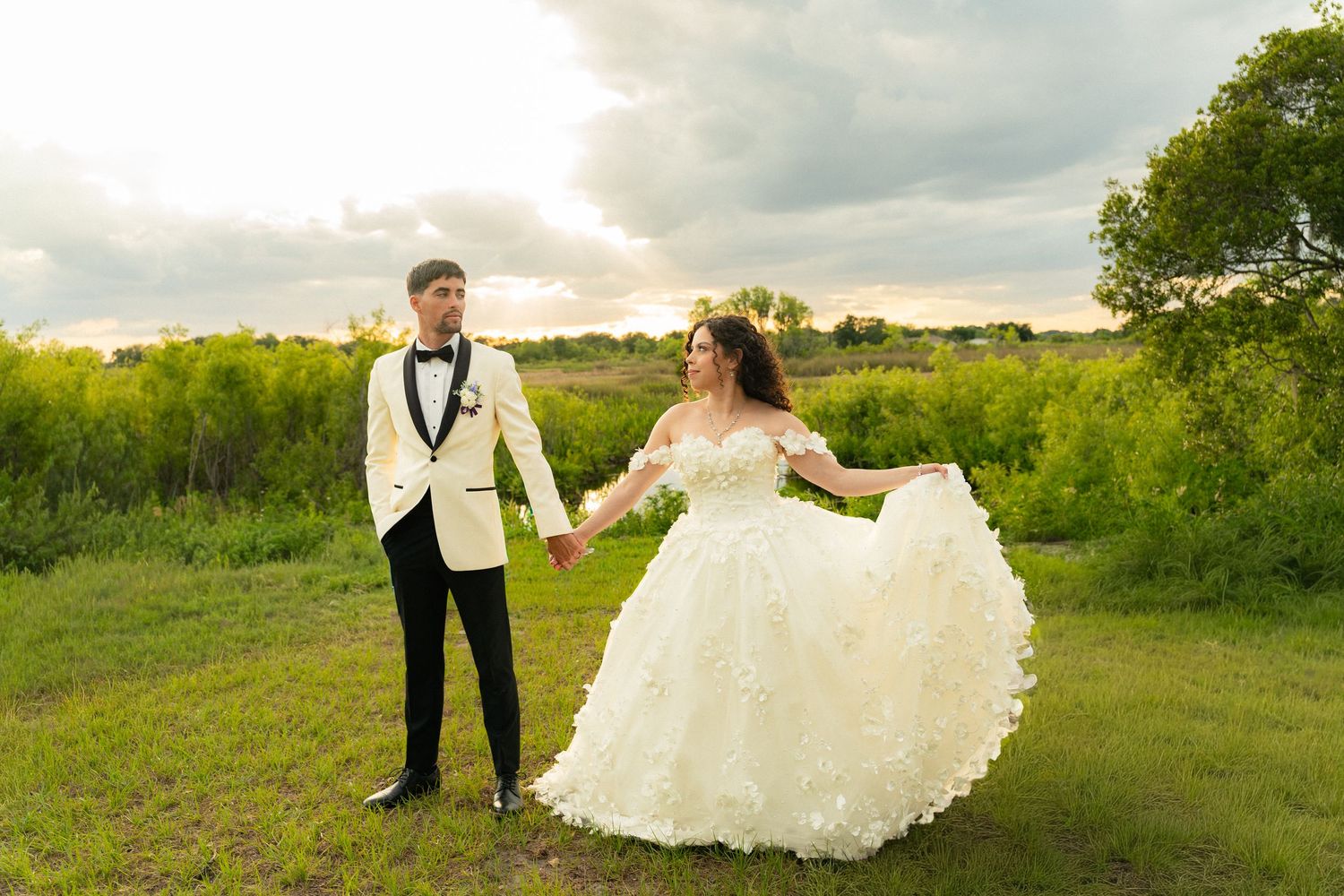 Bride twirling ruffled ball gown while holding grooms hand in lush green field at sunset.