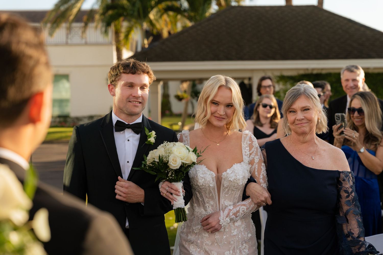 Bride in lace gown holding white bouquet walks down aisle with groom in tuxedo and mother at outdoor wedding.