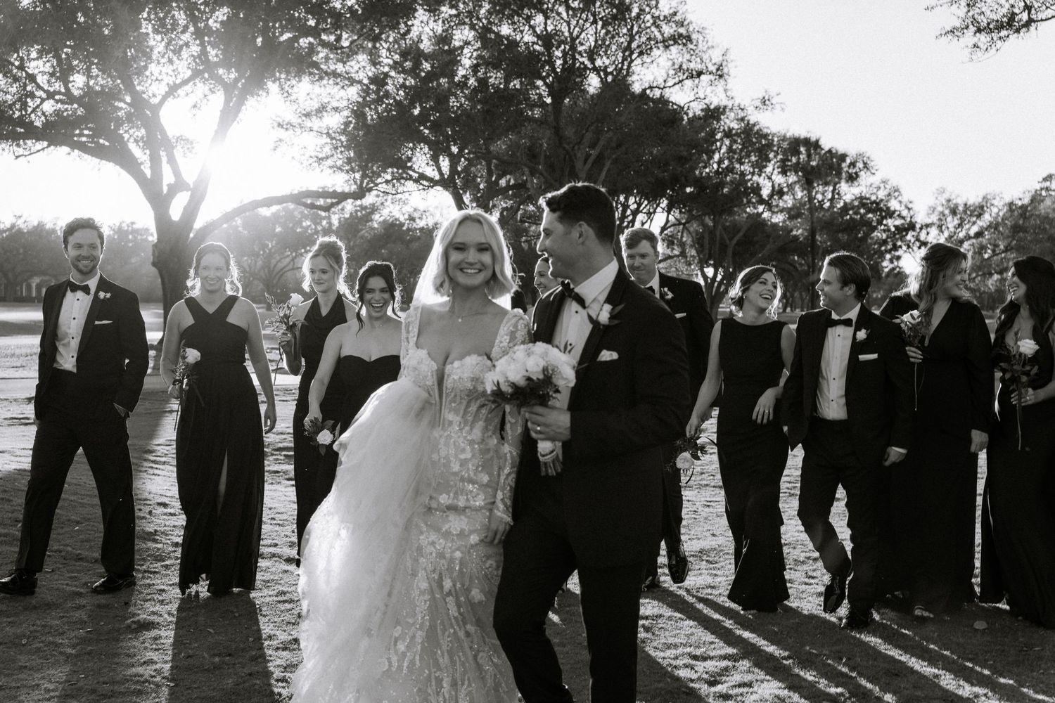 Black and white photo of a bride and groom walking with their wedding party outdoors under trees.