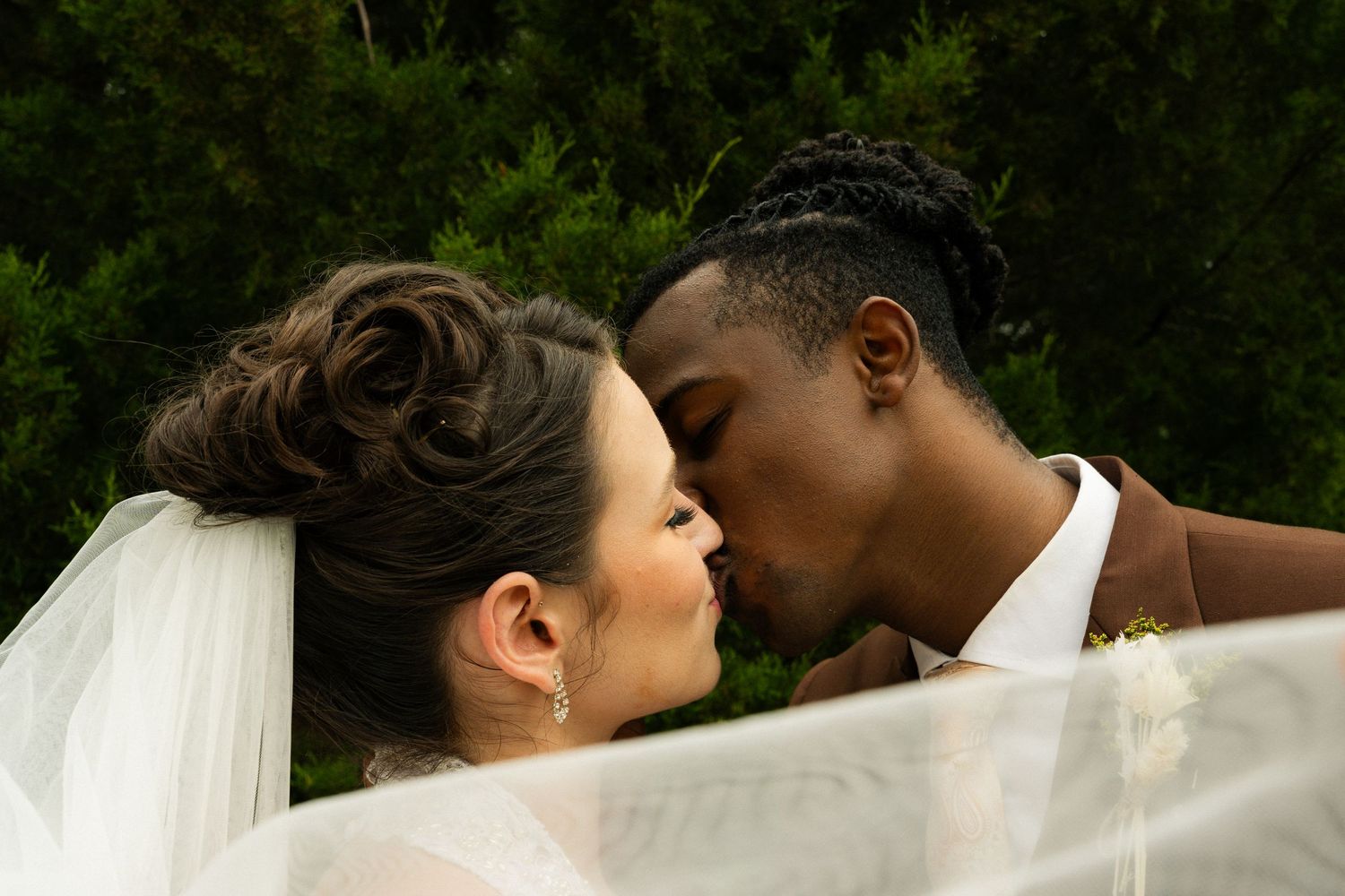 Interracial couple sharing a romantic kiss on their wedding day, bride in veil and groom in brown suit.