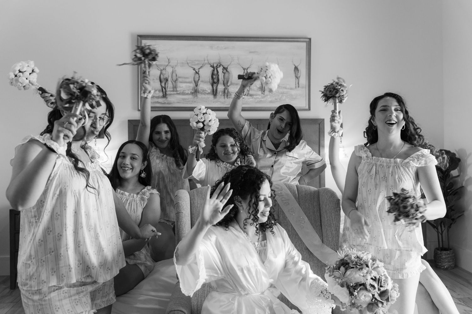 Black and white photo of bride and bridesmaids in robes joyfully raising bouquets during wedding preparations.