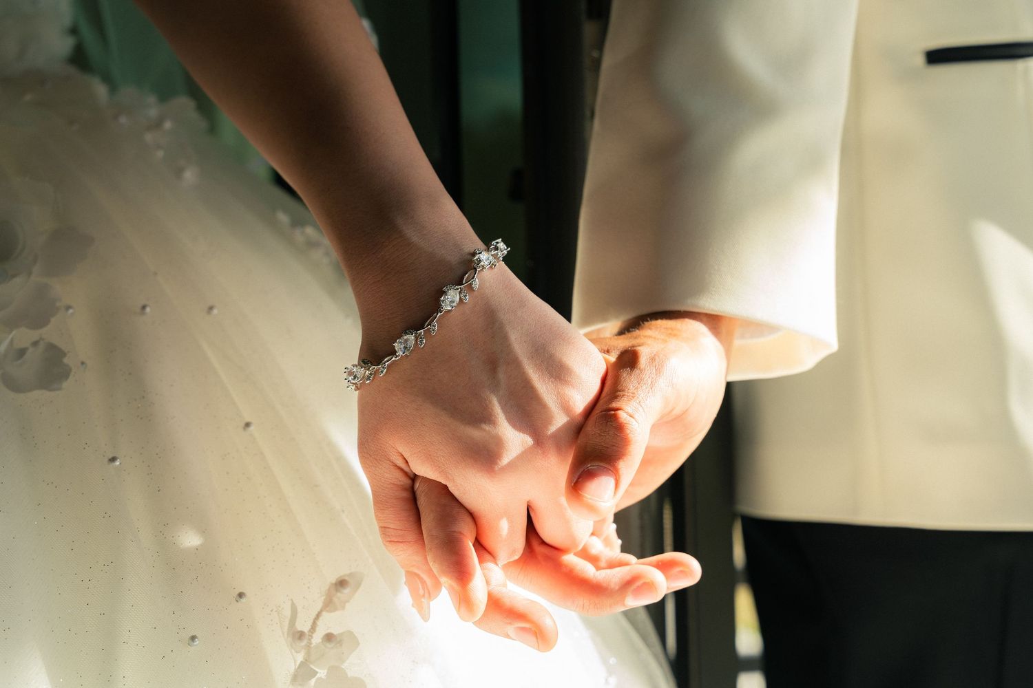 Close-up of bride and groom holding hands showing diamond bracelet and wedding attire details.