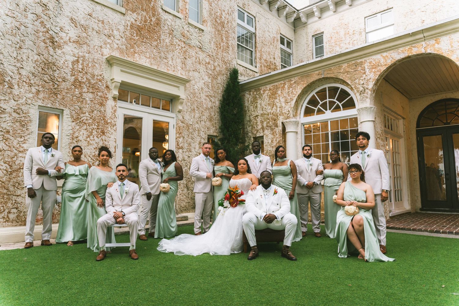 Wedding party poses outside elegant stone mansion, bride and groom seated center, bridesmaids in sage green, groomsmen in gray suits.