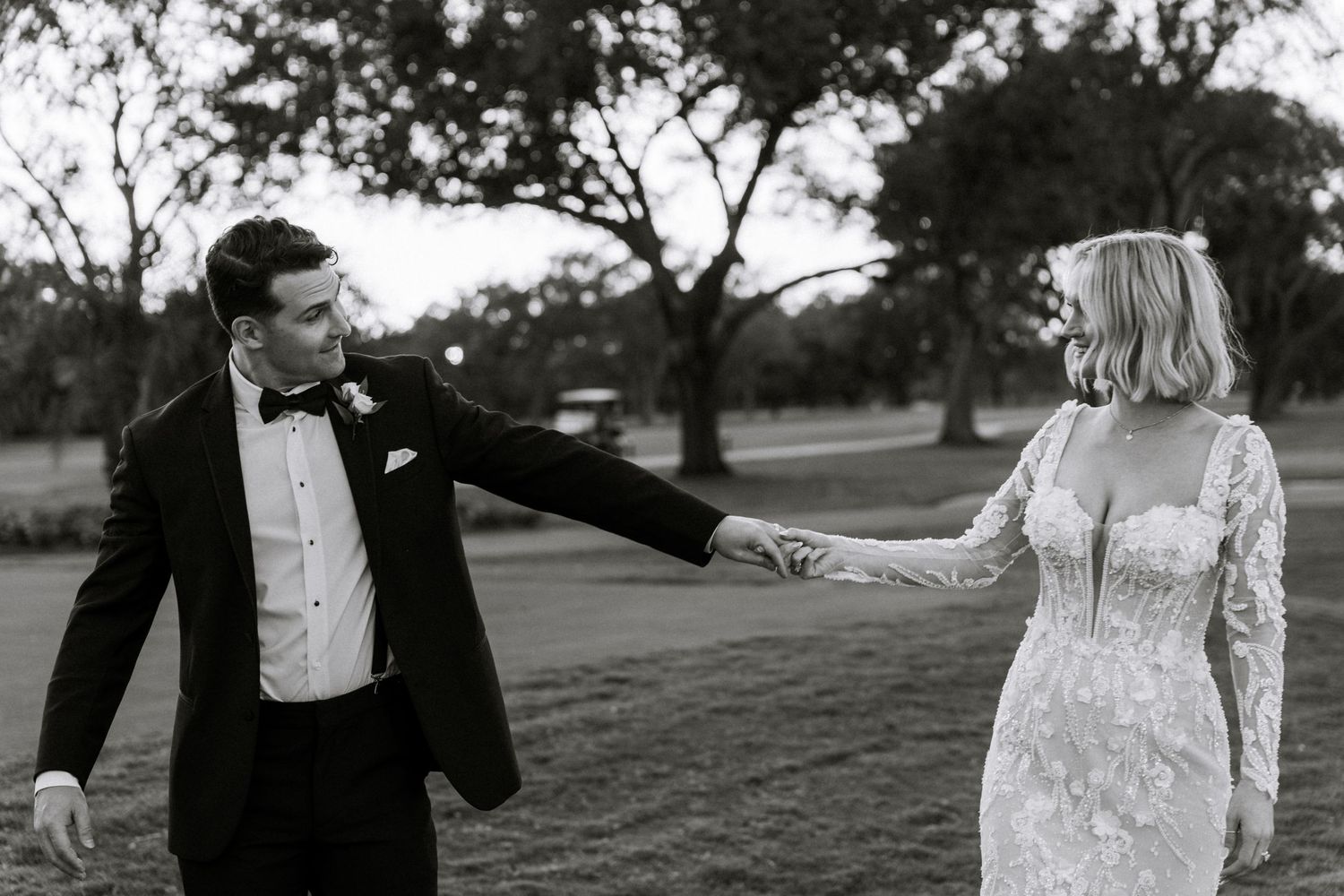 Black and white photo of bride and groom holding hands on golf course in elegant wedding attire.