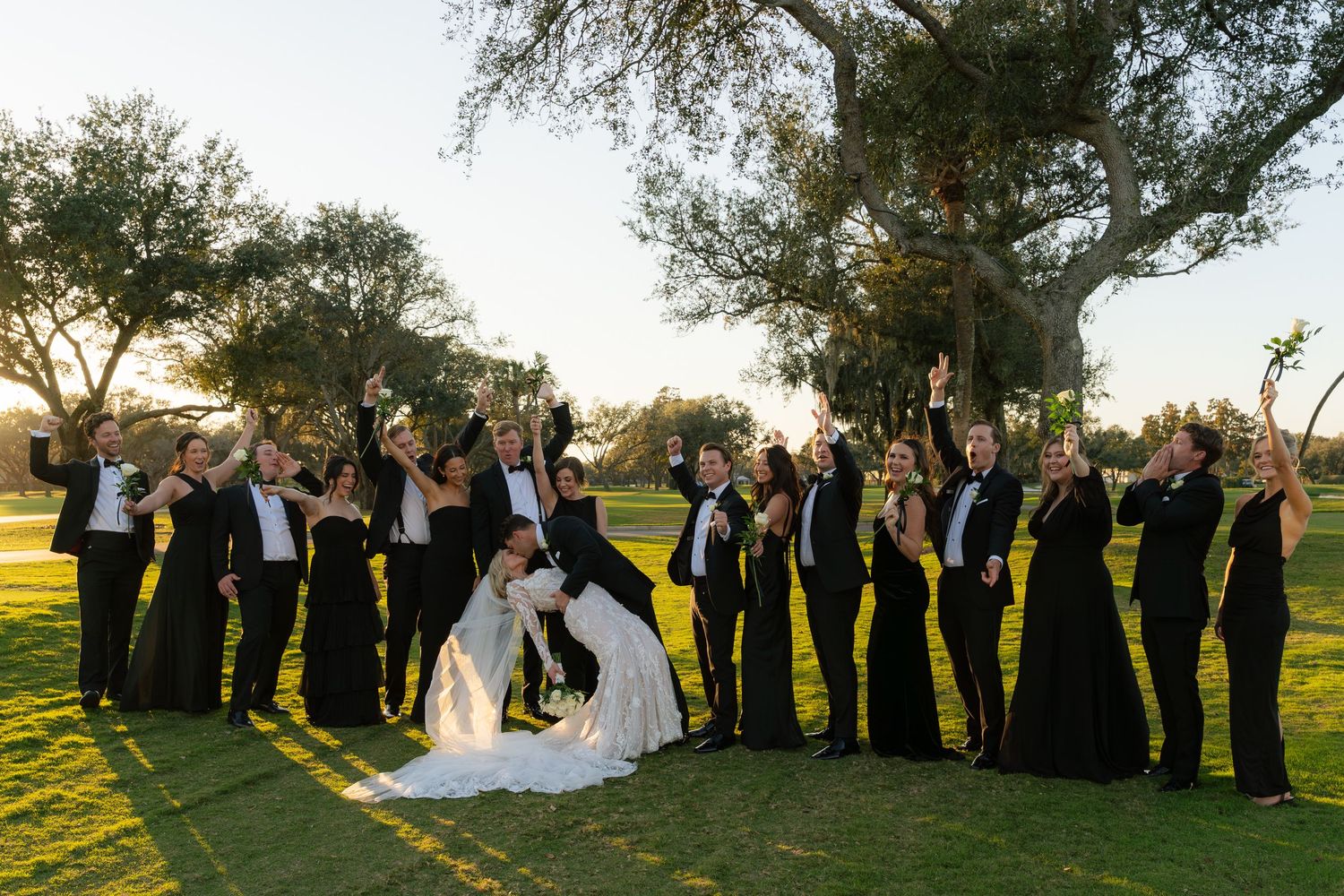 Wedding party in black attire cheers outdoors under oak trees on a golf course as bride and groom share a romantic dip kiss.