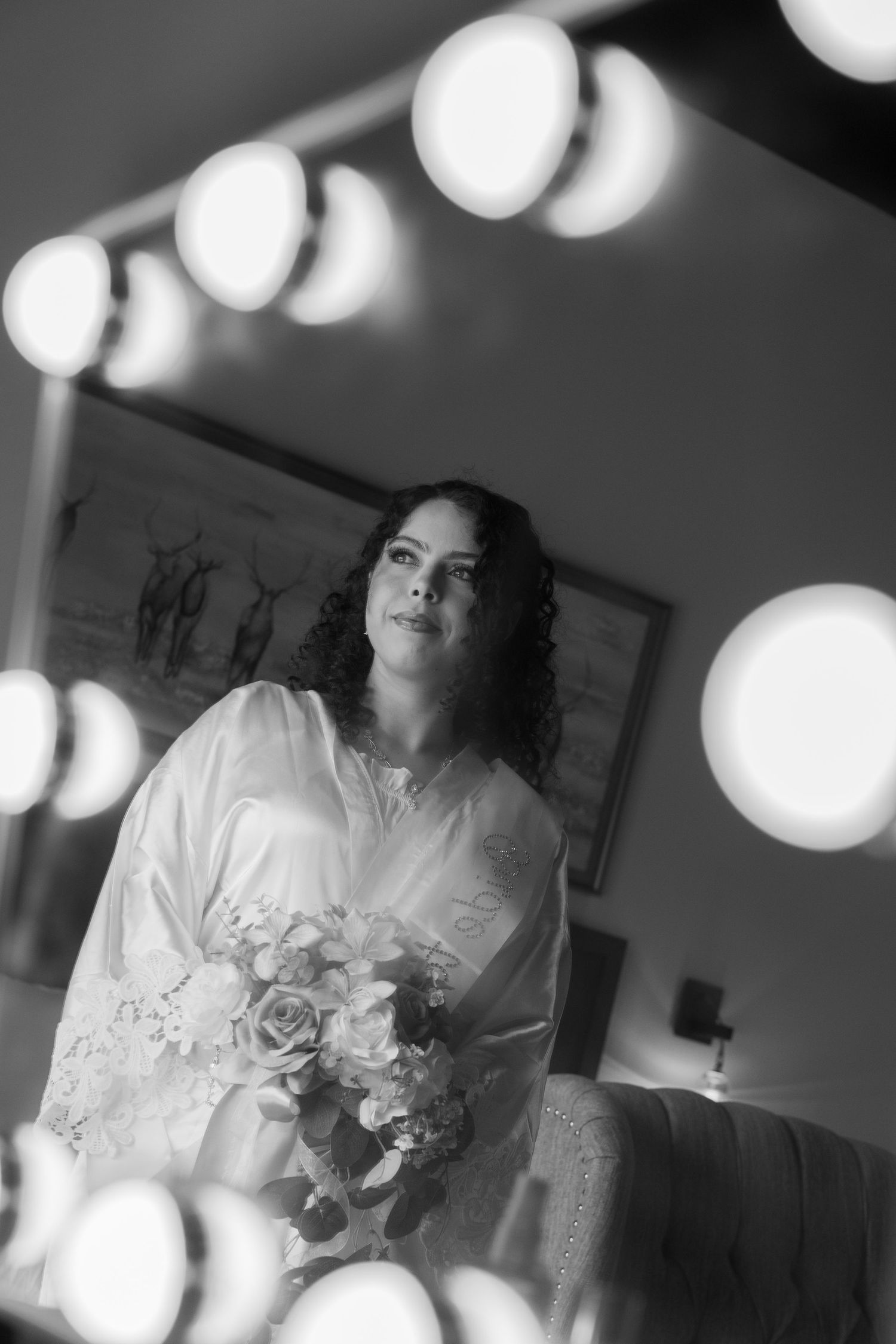 Black and white photo of a bride holding a bouquet reflected in a Hollywood vanity mirror with glowing bulbs.
