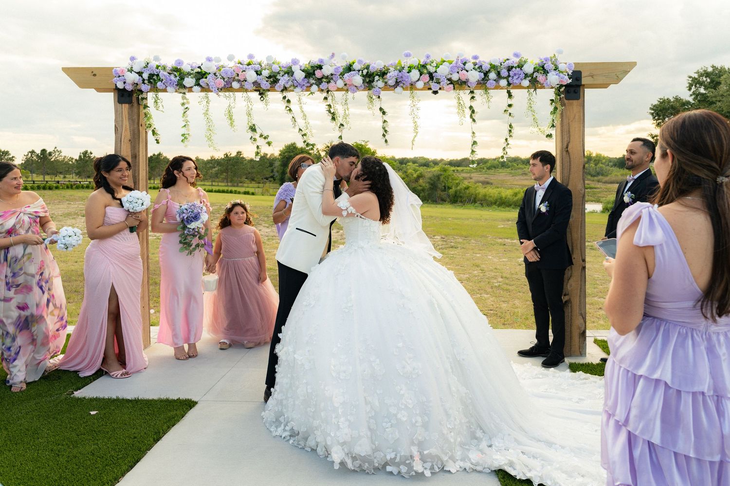 Bride and groom share a kiss under a floral arch surrounded by bridesmaids in pink dresses at an outdoor wedding.