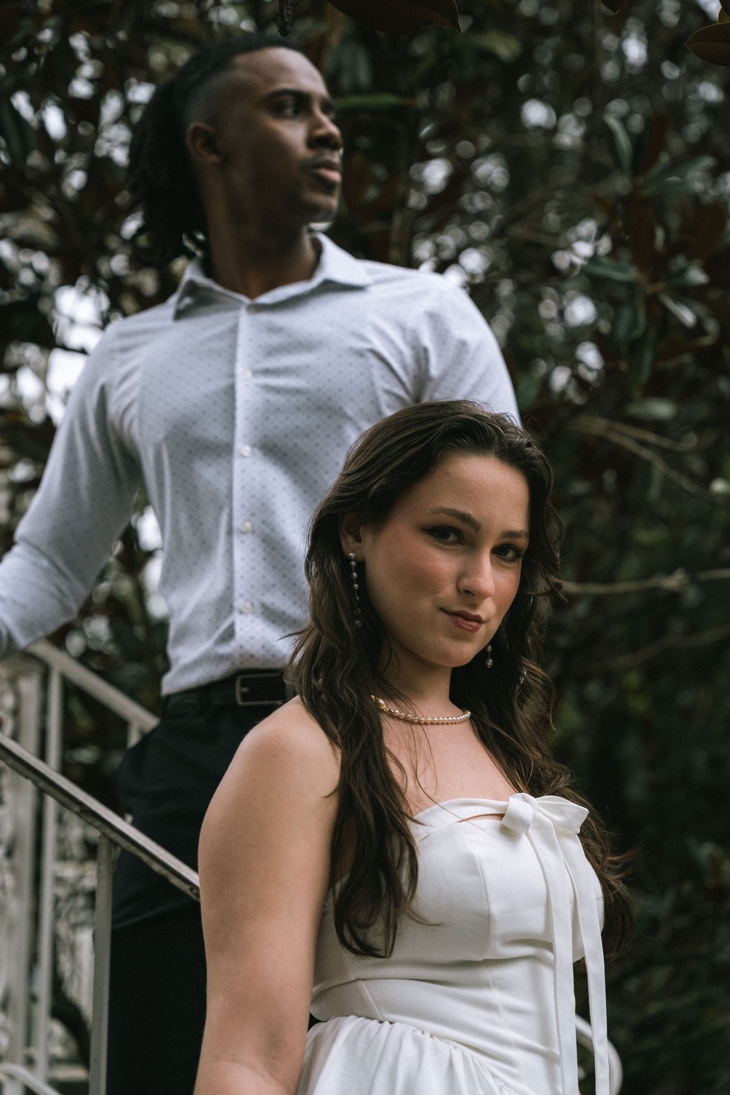 A couple poses on outdoor steps surrounded by green foliage, with one standing and one seated in elegant attire.