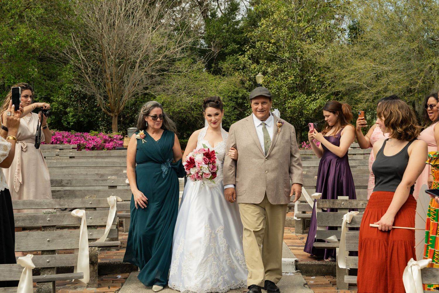 Bride walks down outdoor aisle with parents, guests cheering, lush greenery and pink flowers in background.