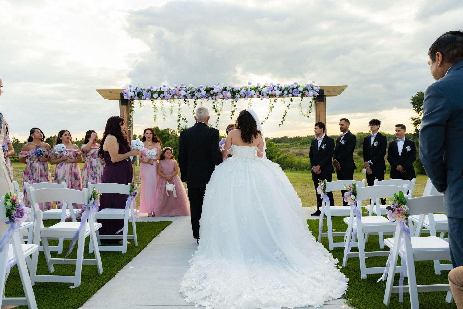 Bride walks down the aisle with escort at outdoor wedding ceremony with purple floral arch and guests.
