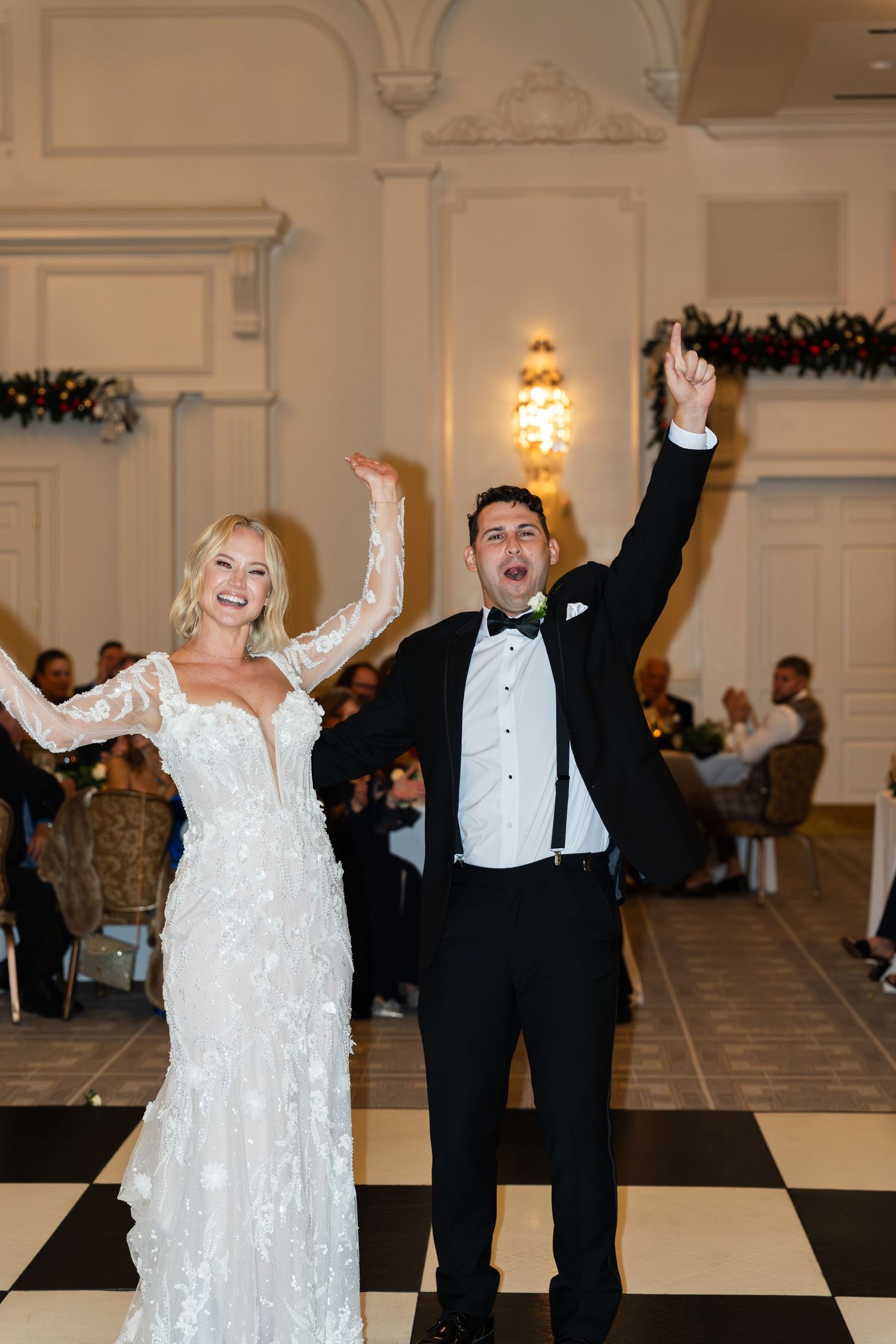 Joyful bride and groom raise arms celebrating grand entrance at elegant ballroom wedding reception.