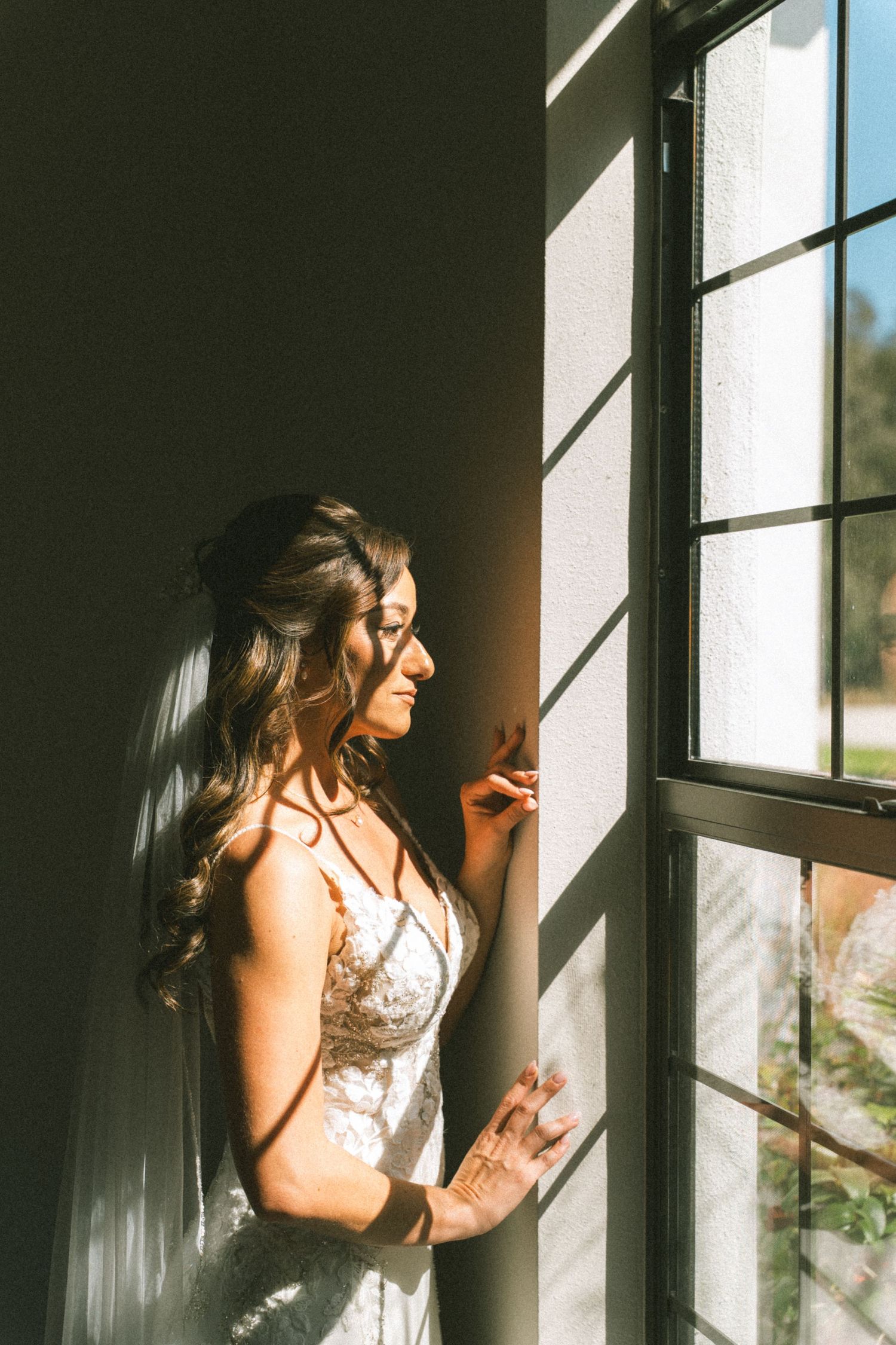 Bride in lace wedding dress standing by sunlit window with flowing hair and veil, bathed in warm natural light.