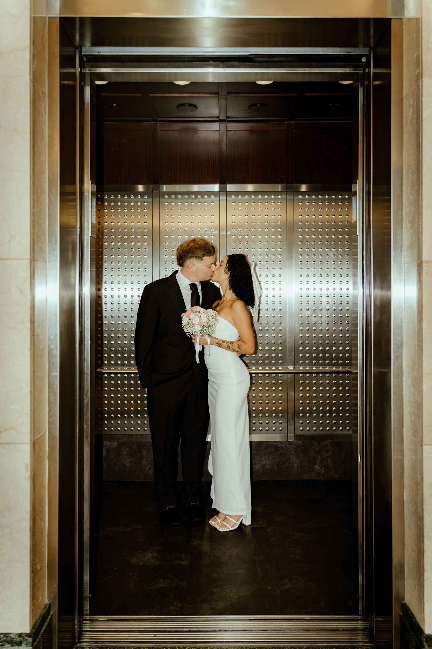 Couple sharing a kiss in a gold elevator, bride in white gown holding bouquet, groom in black suit.