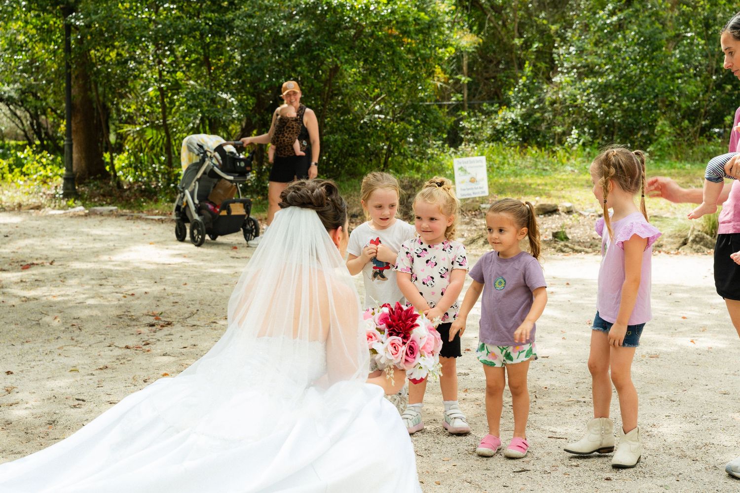 Bride in white gown interacting with young children outdoors at a wedding celebration in a park setting.