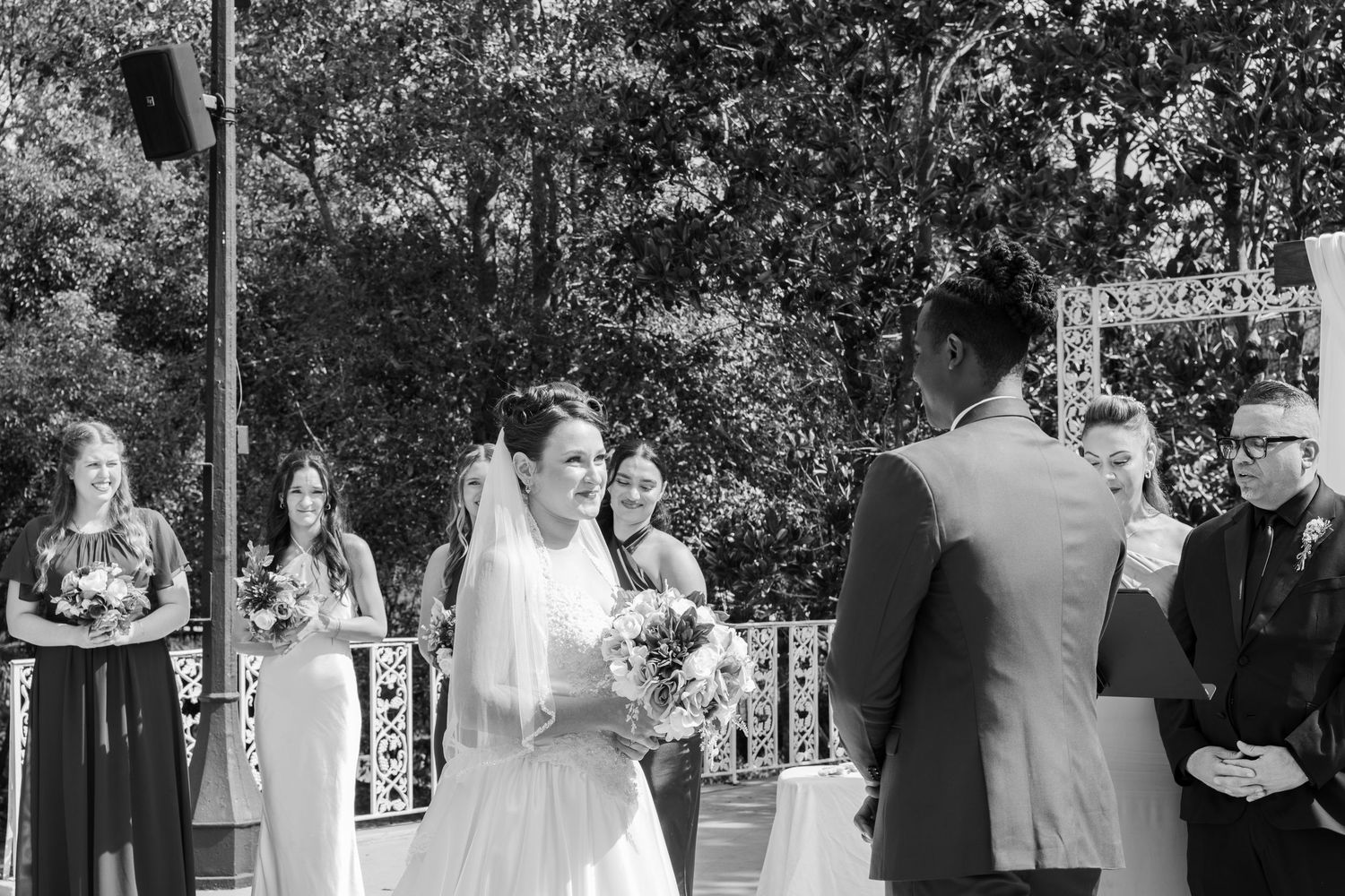 Bride smiles at groom during outdoor wedding ceremony, bridesmaids and guests standing nearby.