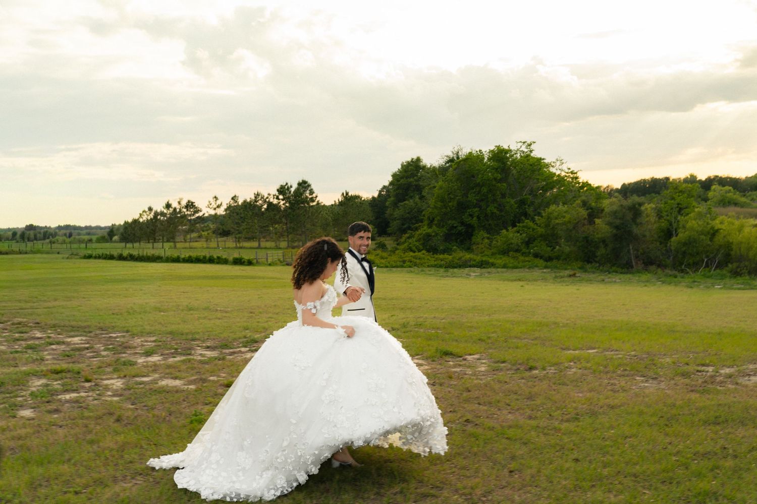Groom lifts bride in stunning white ball gown in a lush green field at sunset.