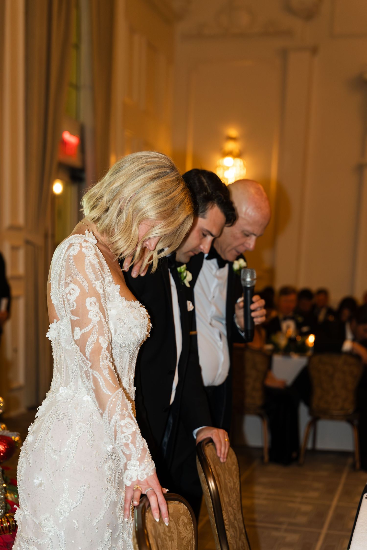 Bride in lace gown and groom bow heads during wedding reception toast inside elegant ballroom.